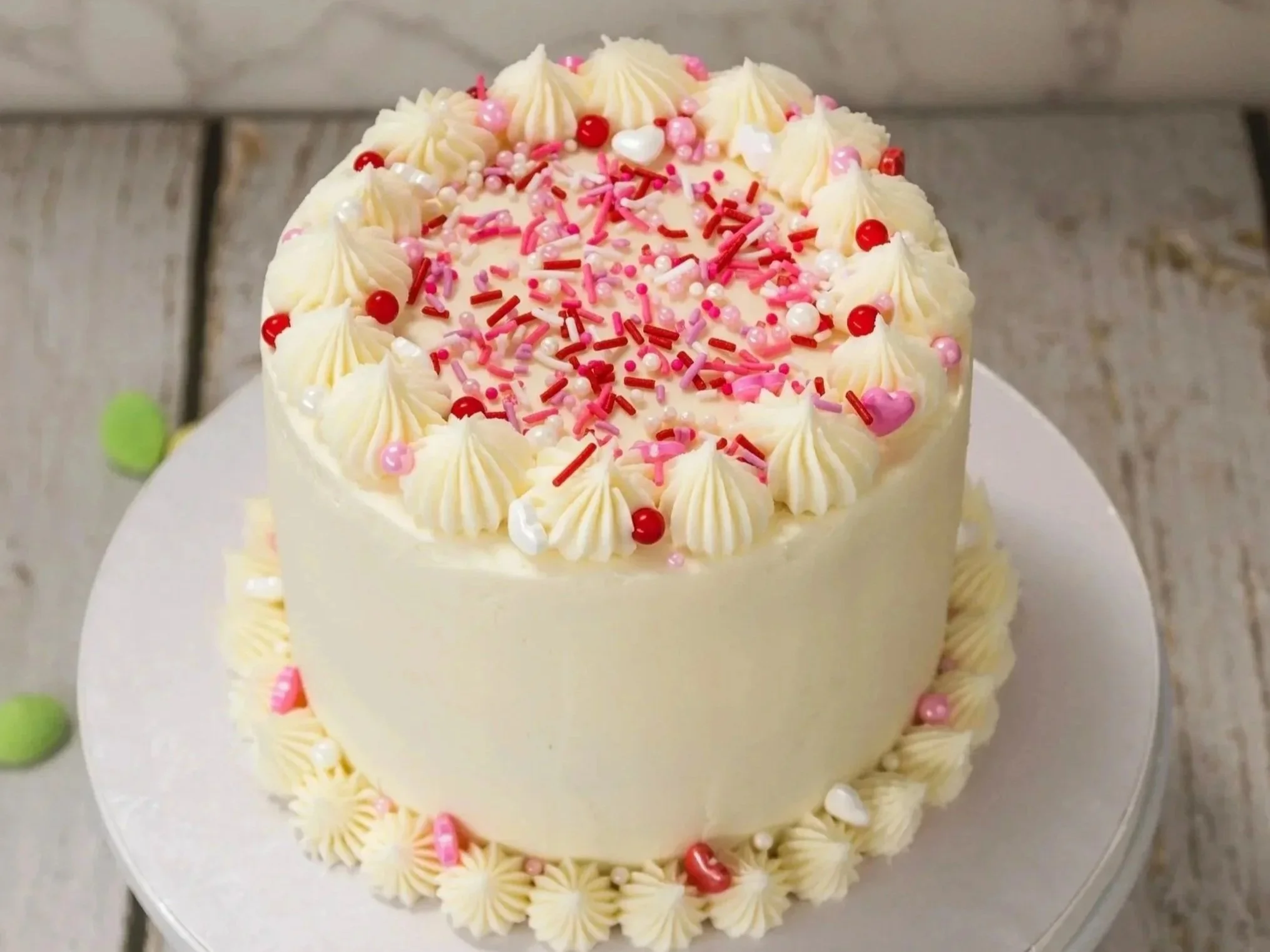A white birthday cake decorated with pink, red, and white sprinkles, with piped cream rosettes around the top edge, on a white cake stand.