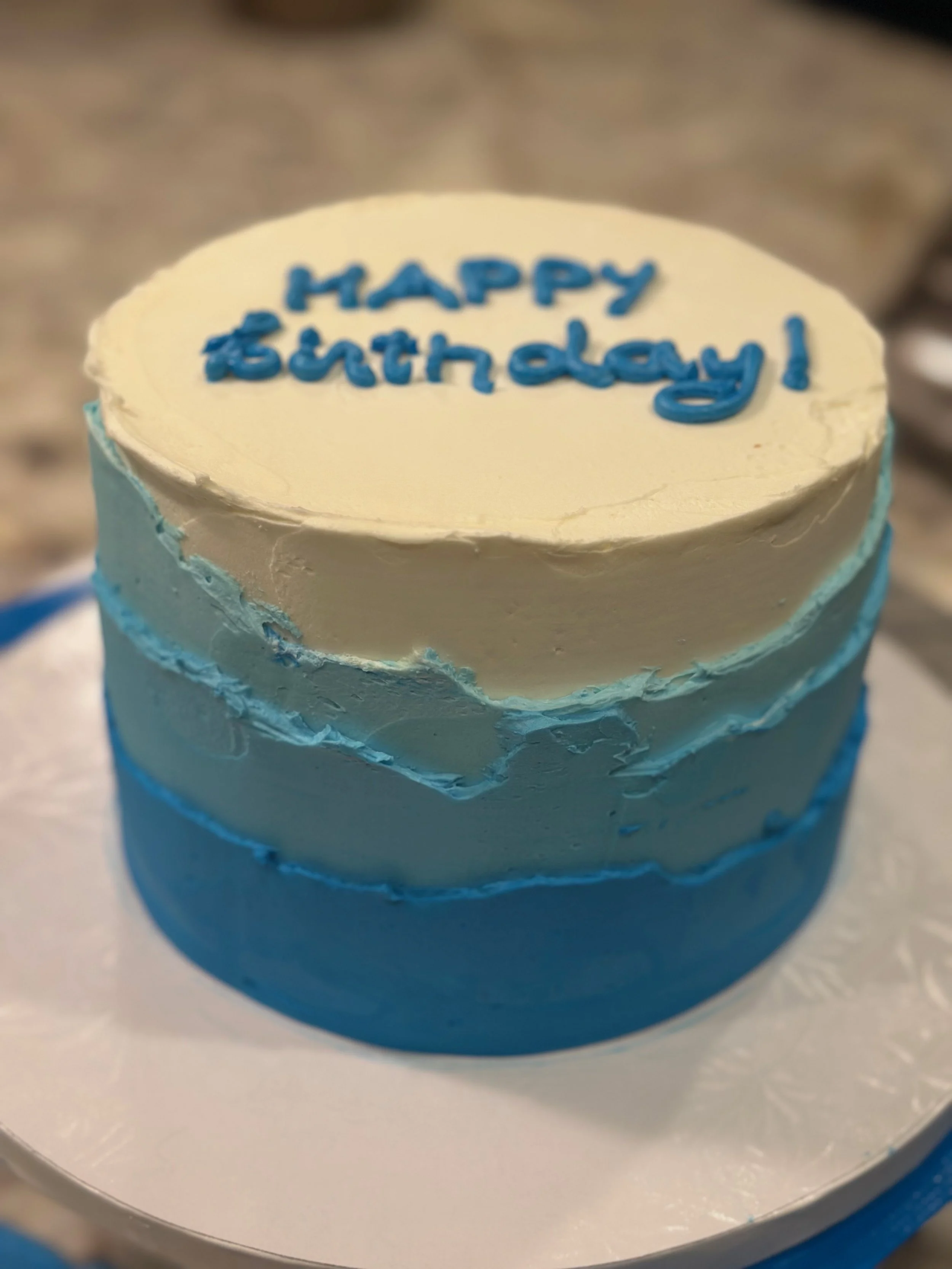 A round cake with blue and white frosting, with a message that reads 'Happy Birthday!' in blue icing.