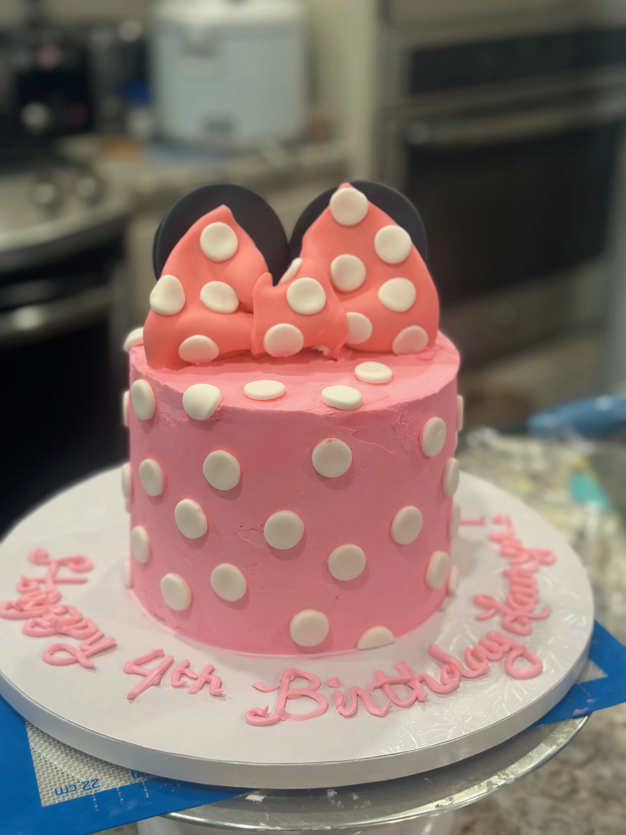 Pink birthday cake decorated with white polka dots and pink icing, topped with two pink and black Minnie Mouse shaped fondant decorations, with a 'Happy Birthday' message written in pink on the cake board.