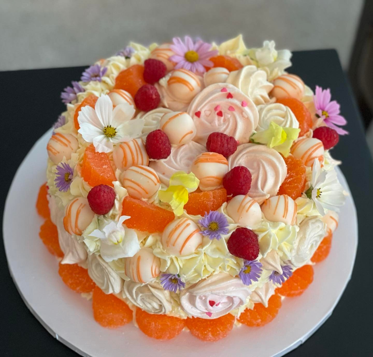 Decorated cake with white, pink, and orange frosting, fresh raspberries, mandarin orange slices, edible flowers, and colorful sprinkles.