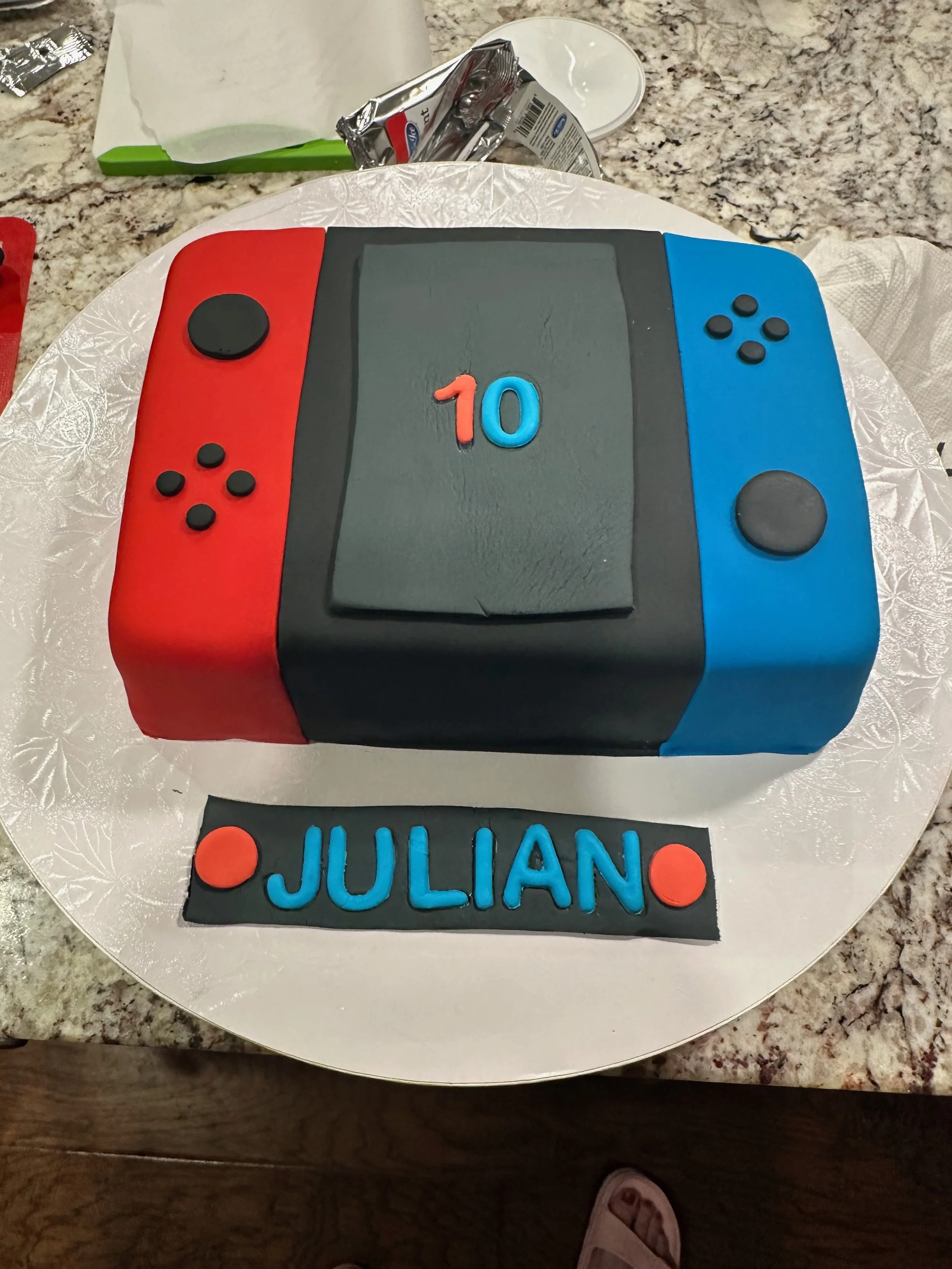 A cake decorated to look like a Nintendo Switch gaming console, with red and blue controllers on each side and the number 10 in the middle. The cake also has the name 'Julian' on a black fondant plaque at the bottom.