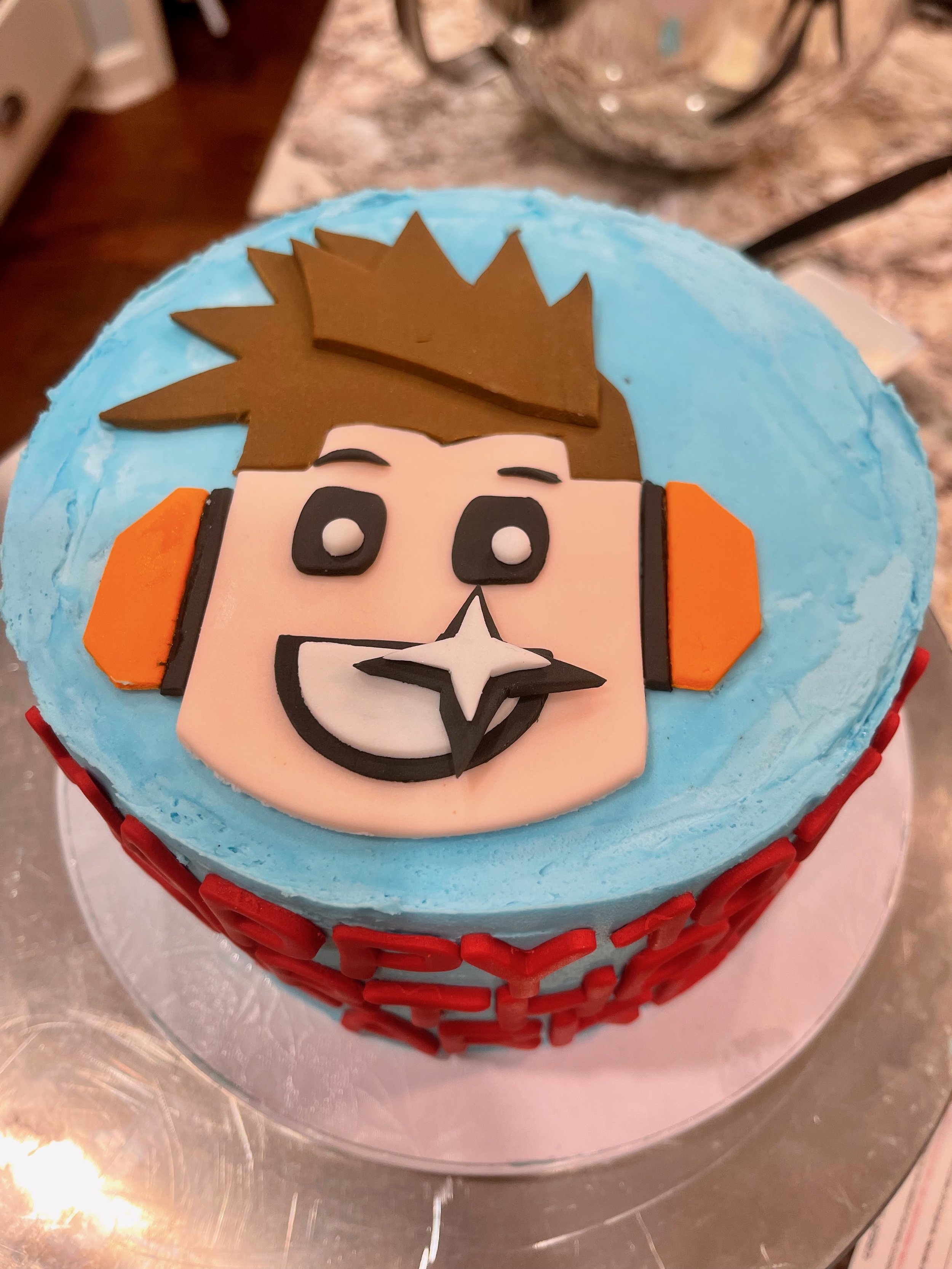 A cake with a face of a cartoon character with spiky brown hair, black eyes, and a big smile, decorated with blue icing on top, orange and black details, and red decorations around the bottom edge.
