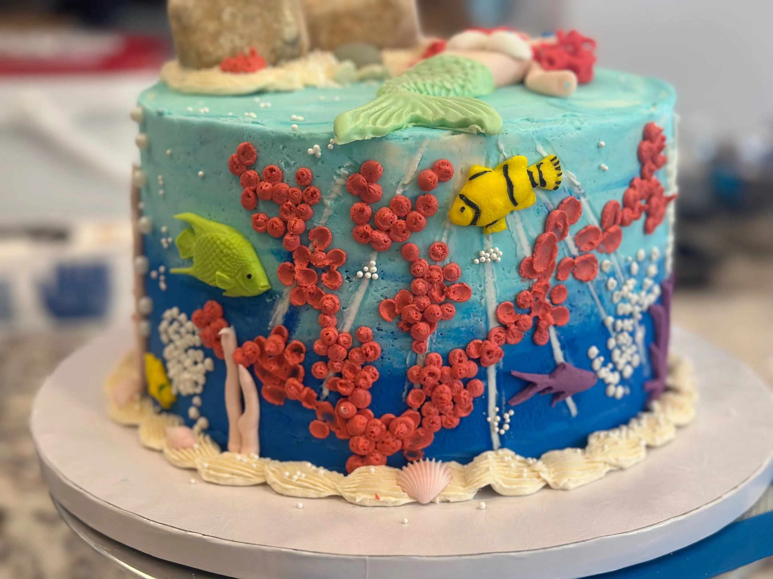 Cake decorated with marine theme; blue icing with red coral-like decorations, green and yellow fish, white seashells, and ocean elements.