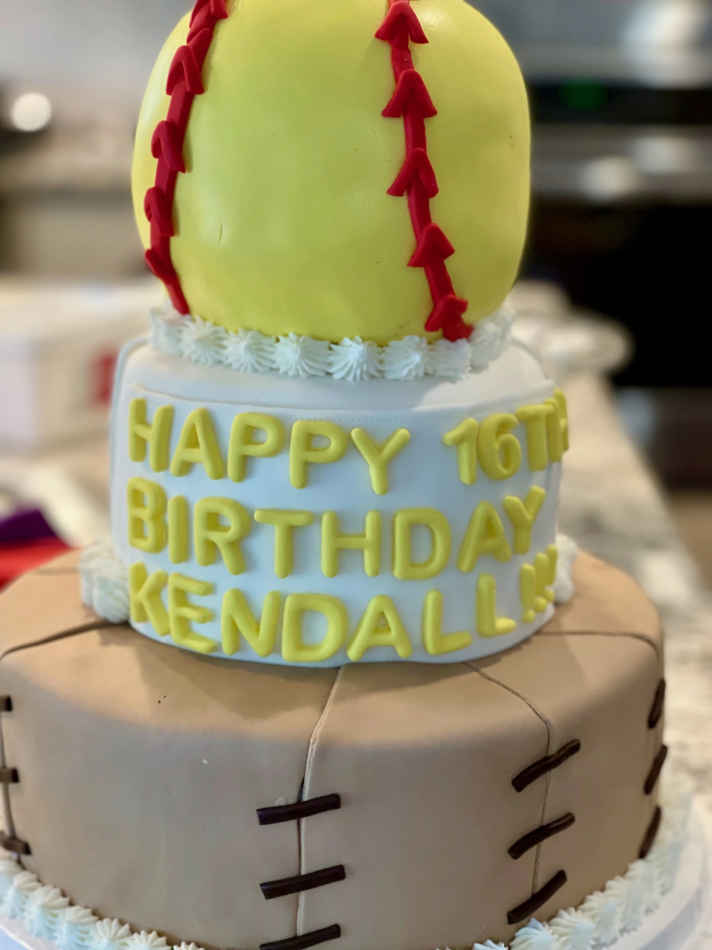 A cake decorated like a softball with a yellow tennis ball on top, featuring red icing stitches. The cake has yellow and black text around it that reads 'Happy 16th Birthday Kendrick.'