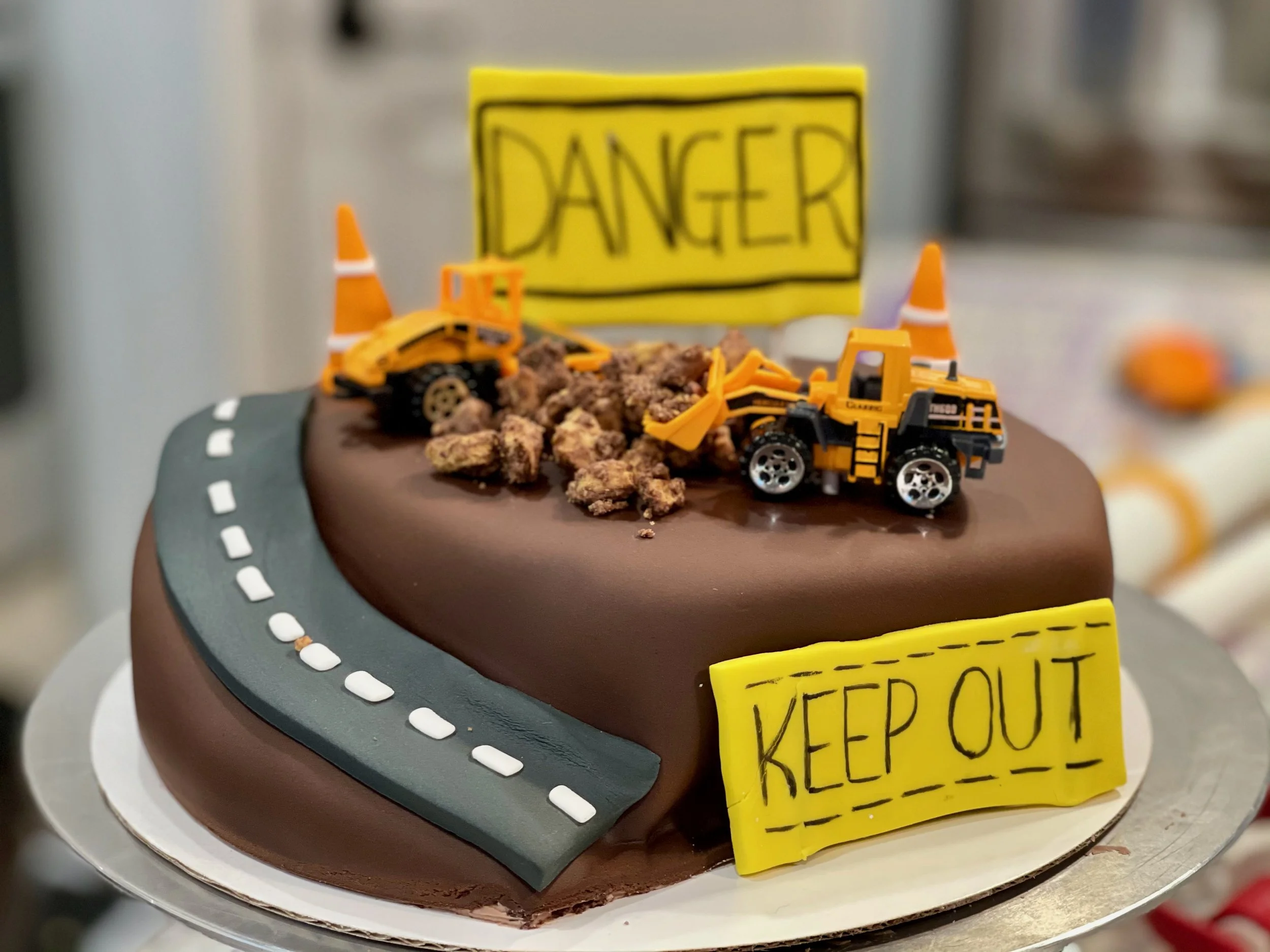 A decorative cake designed to look like a construction site with toy construction vehicles, a road sign, and a caution tape that reads 'KEEP OUT'.