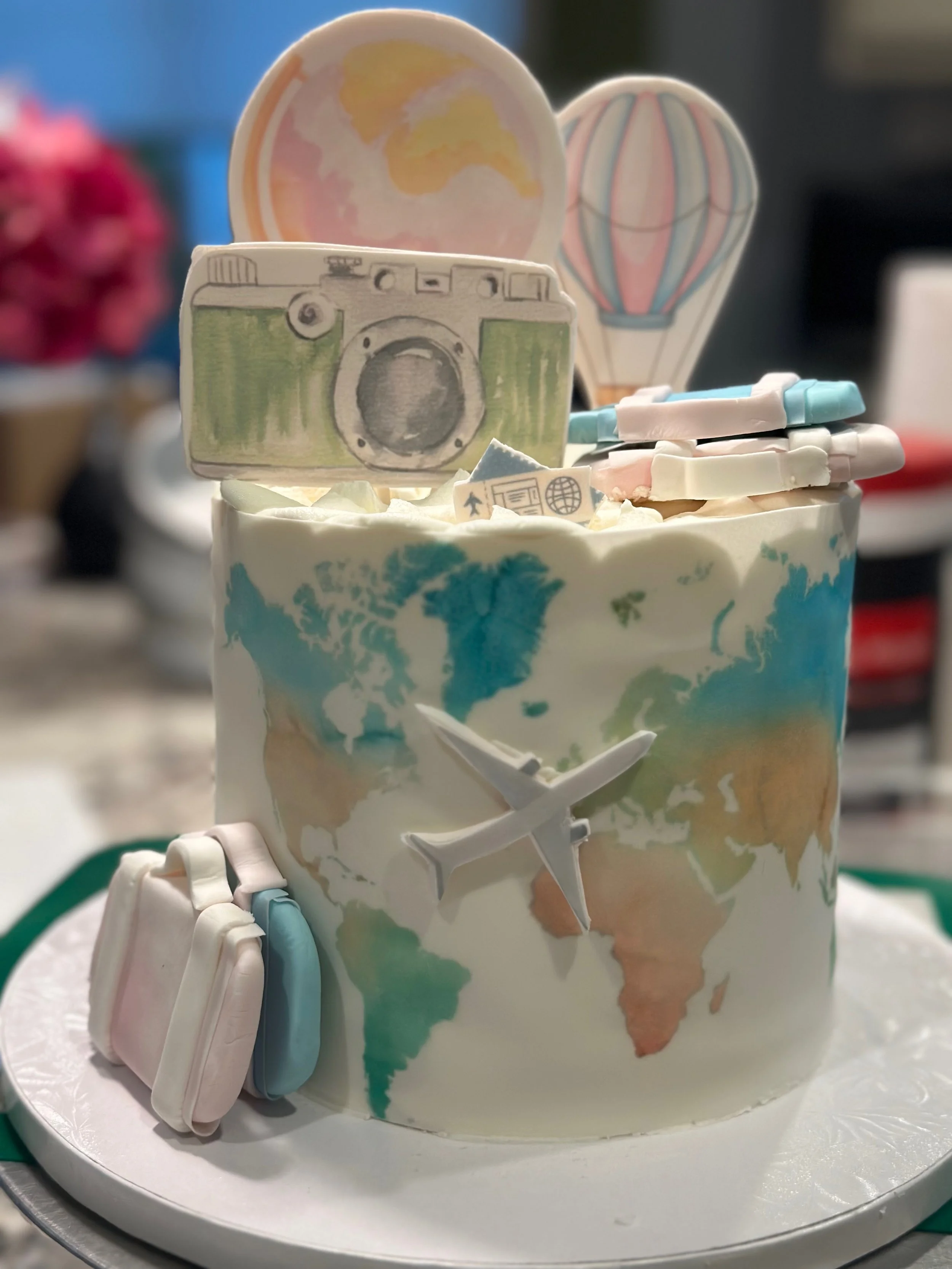 Cake decorated with travel-themed toppers including a camera, an airplane, and hot air balloons, with a world map design on the cake's surface.