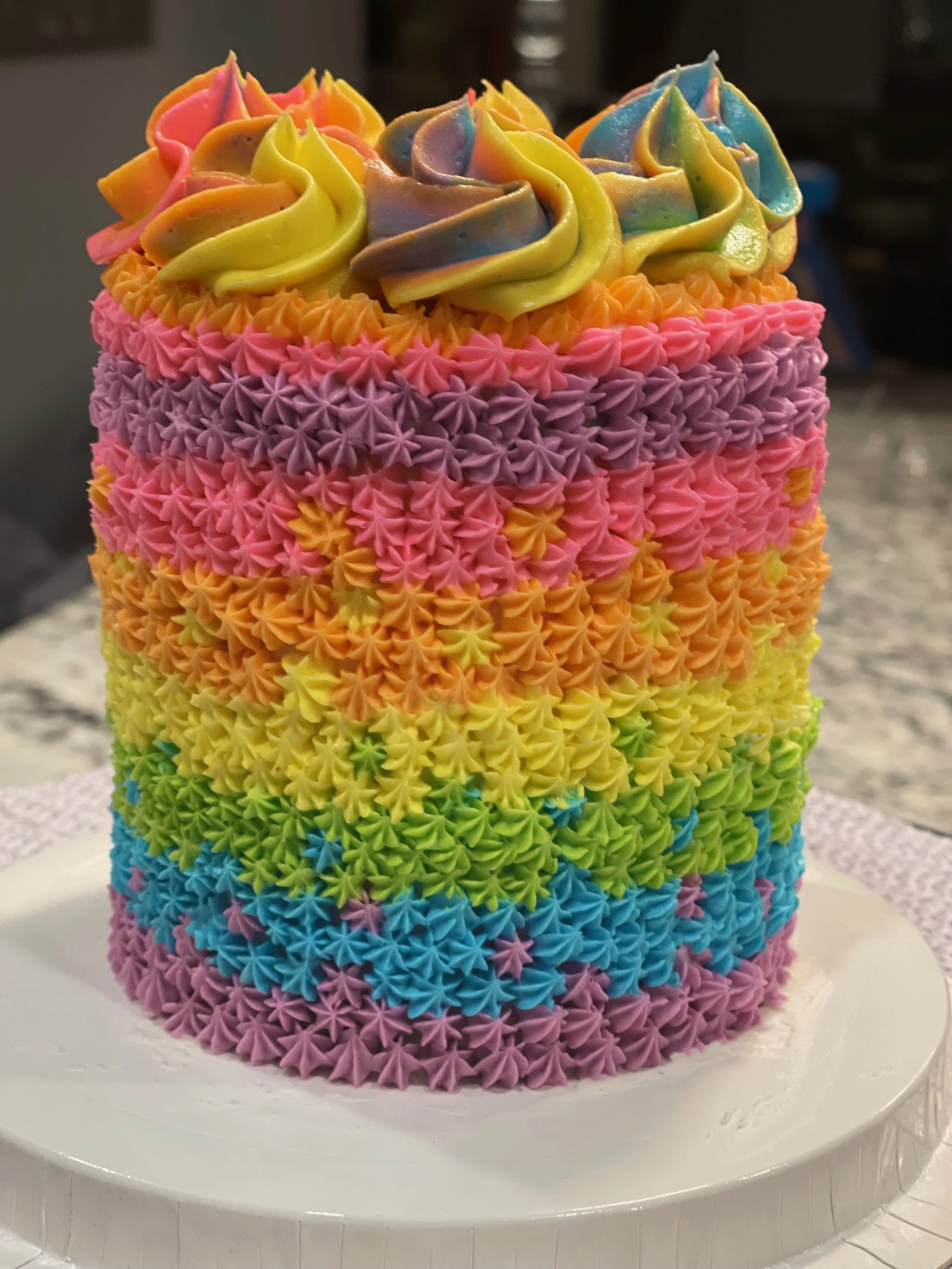 Colorful rainbow cake decorated with multi-colored buttercream frosting in star-shaped designs, stacked in rainbow layers from red at the bottom to purple at the top.