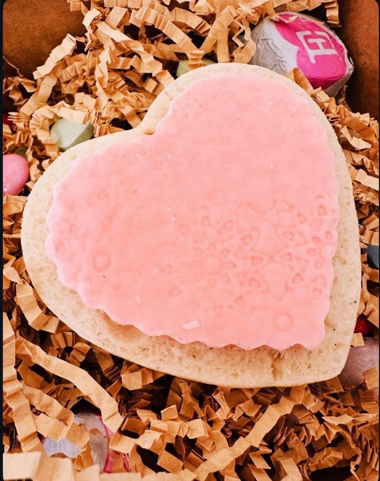 A heart-shaped sugar cookie with pink icing and a decorative pattern, resting on crinkled paper shred inside a gift box with candy and shredded paper.