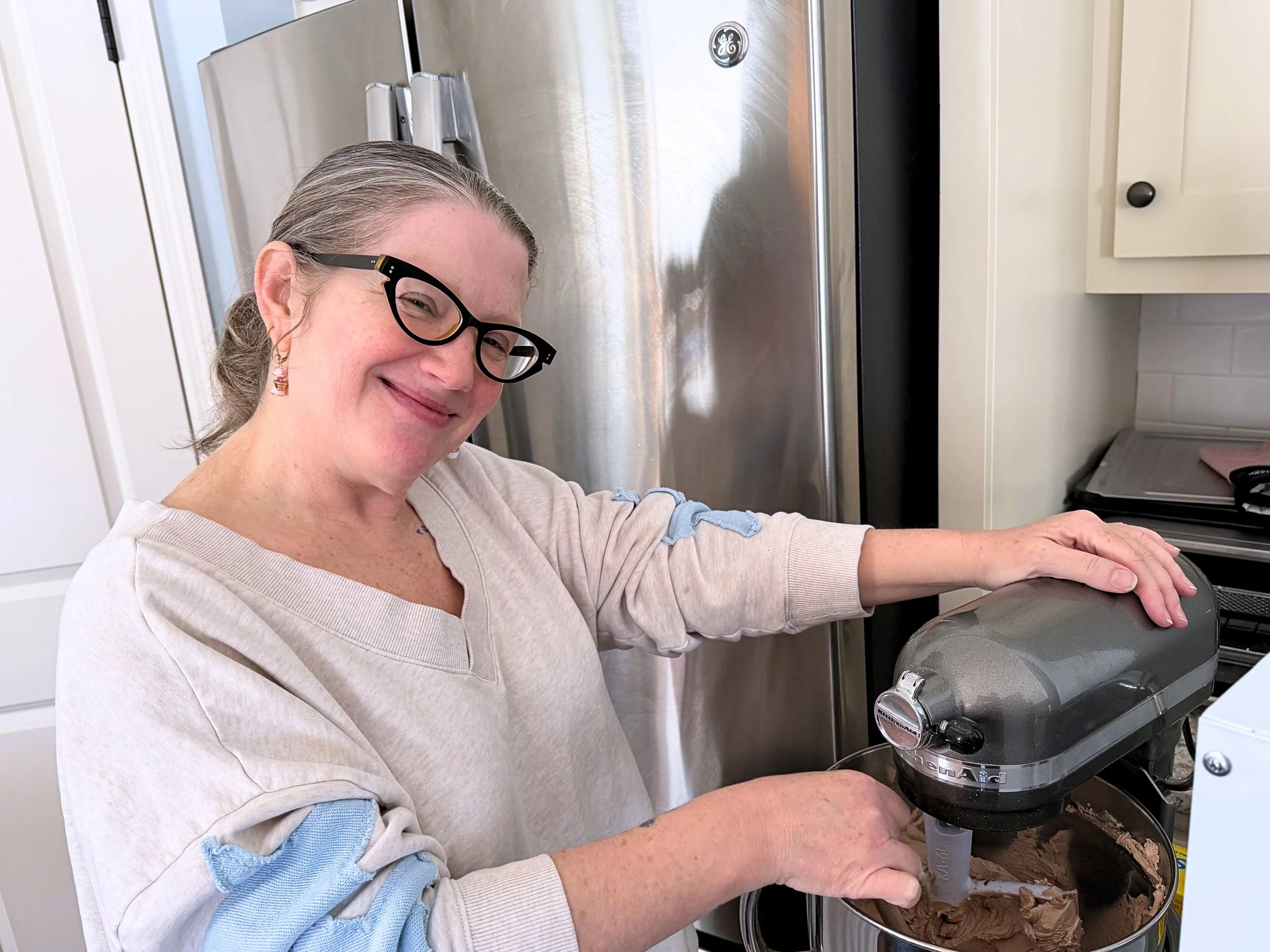 A woman with glasses and earrings stirring cake batter in a Kitchenaid mixer.