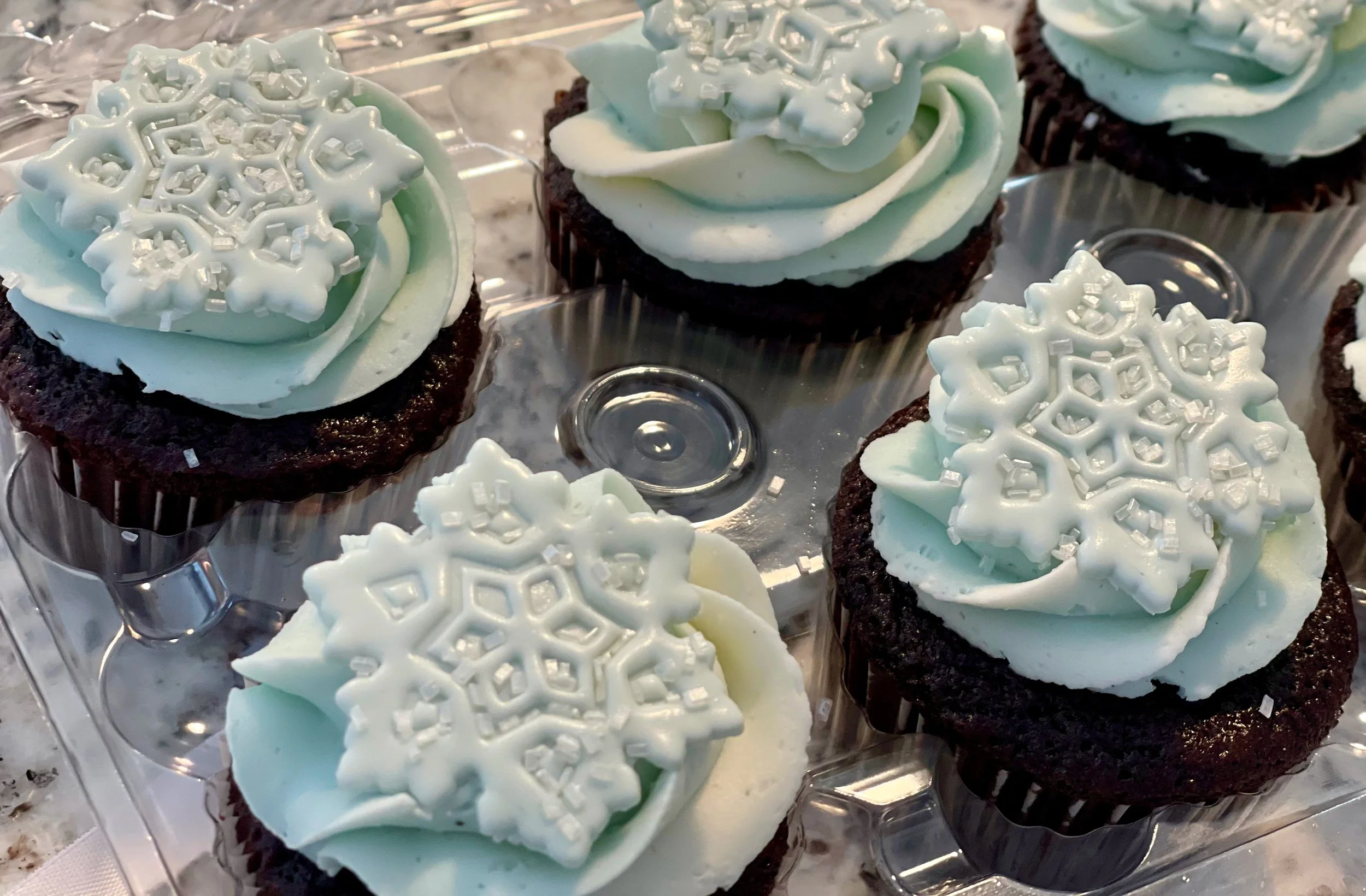 Chocolate cupcakes topped with light greenish frosting and decorated with white snowflake-shaped sprinkles and small silver sugar crystals, arranged in a clear plastic tray.