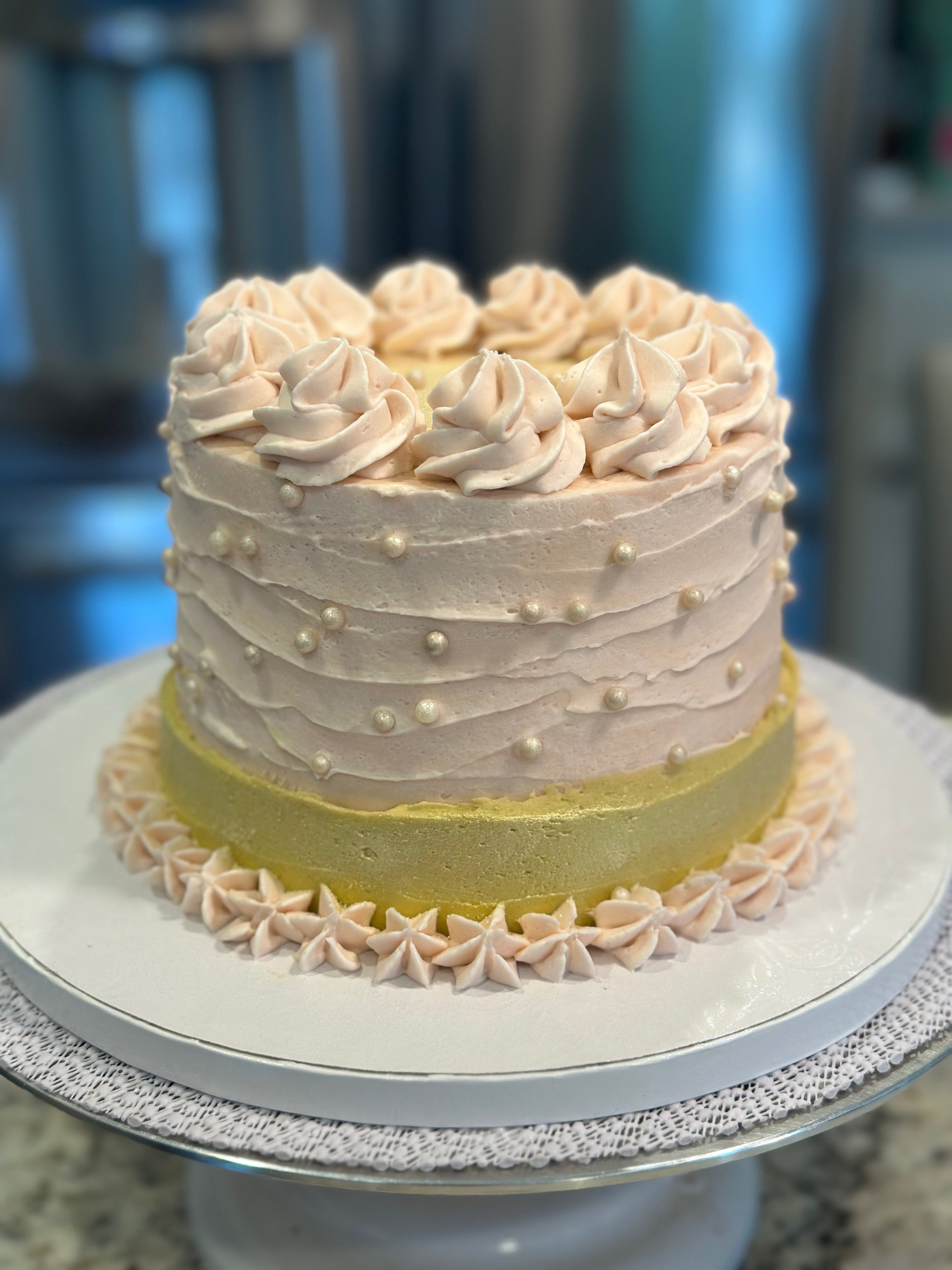 A layered cake with light pink frosting, pearl-like decorations, and piped frosting on top, sitting on a cake stand with a decorative edge.