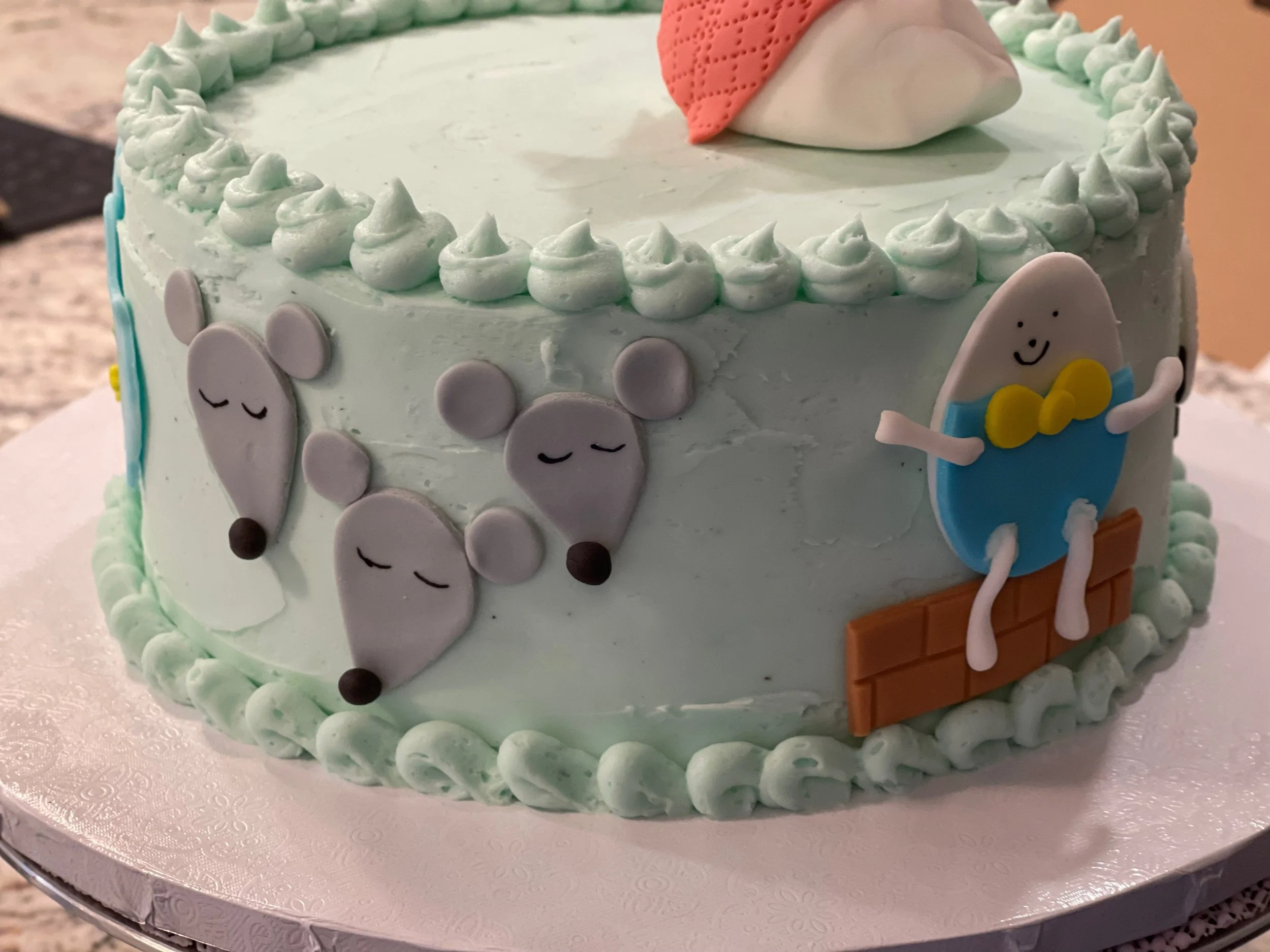 A round birthday cake decorated with light blue icing, featuring cute animal faces and a character in a blue outfit holding a yellow item, surrounded by piped frosting borders.