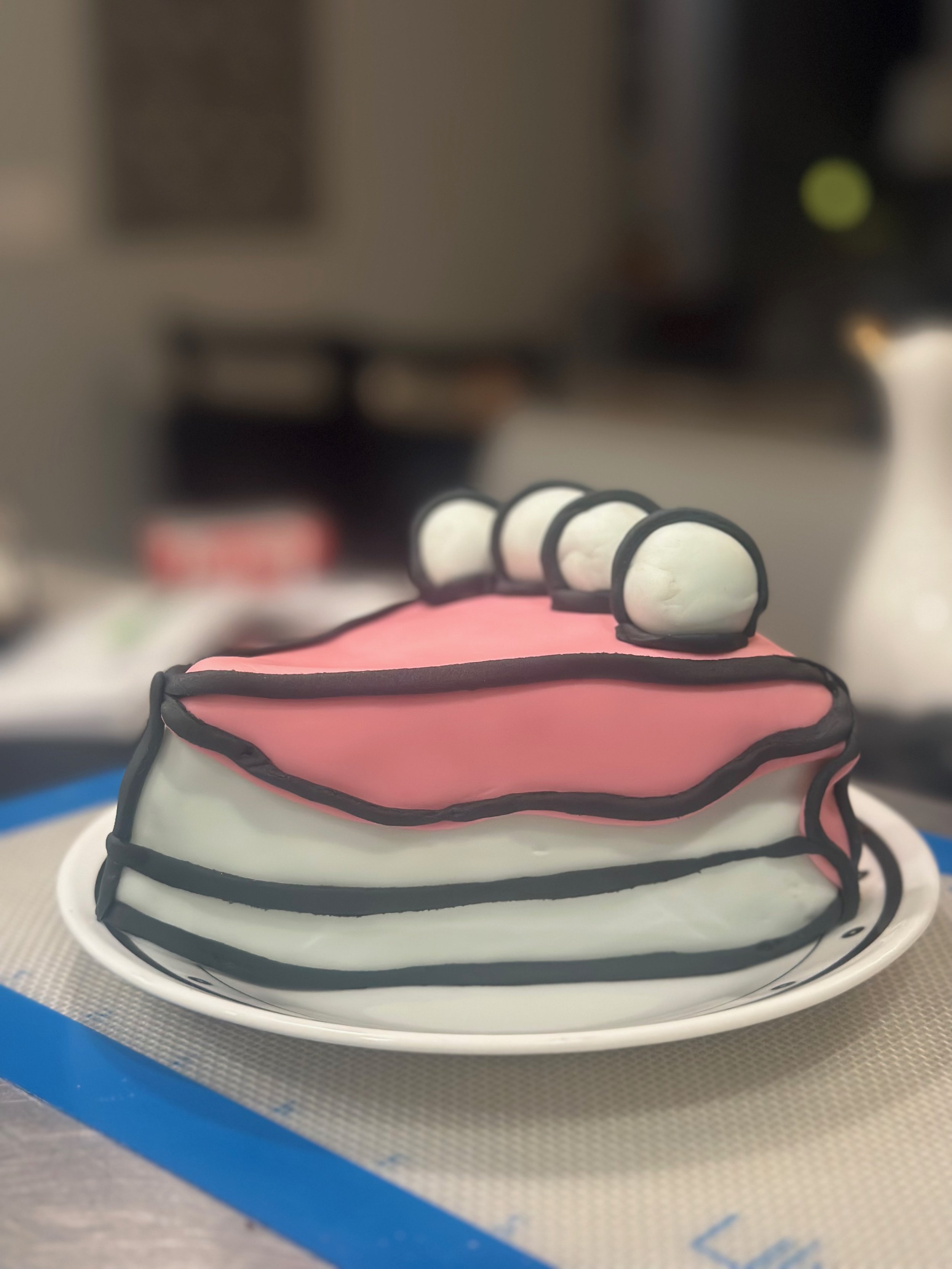 A cake decorated to look like a layered birthday cake with pink, white, and black icing, topped with white fondant balls, sitting on a white plate.