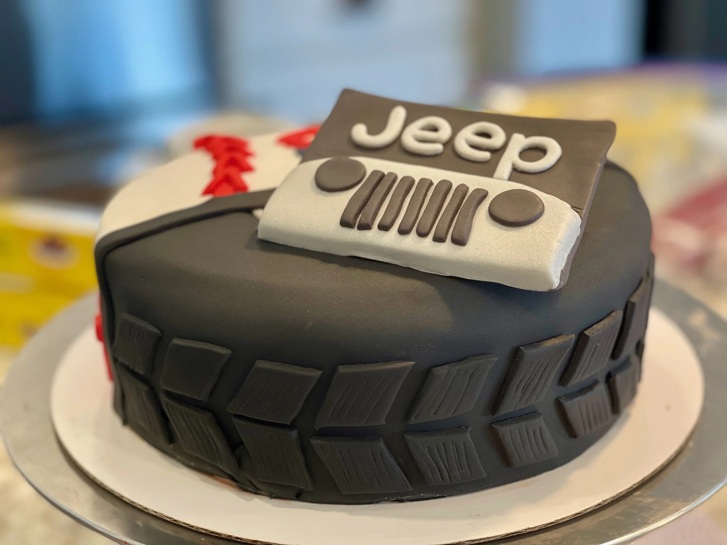A close-up of a tire with a small decorative cake on top, featuring a "Jeep" sign and tire tread design, on a white cake base.
