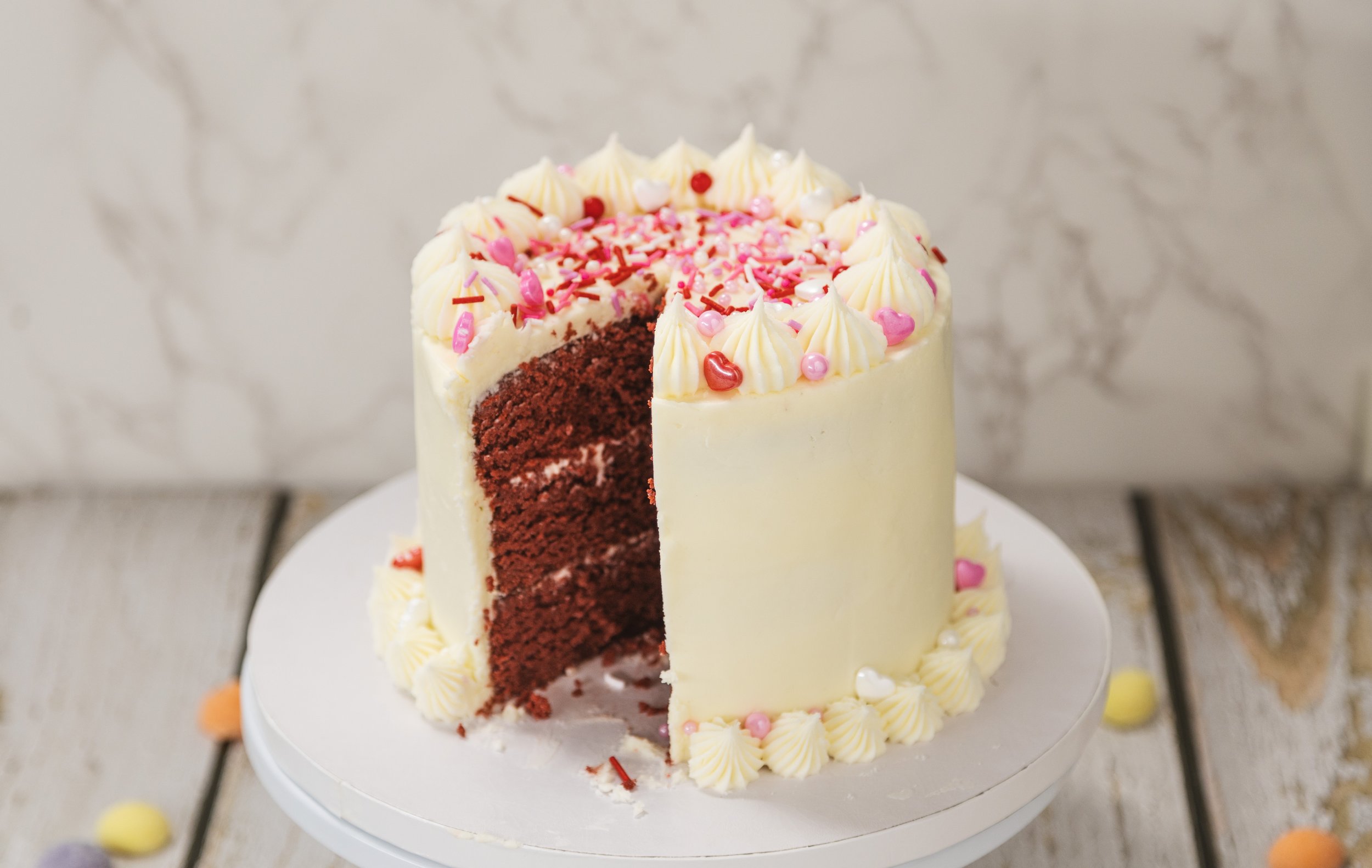 A partially sliced red velvet cake with white frosting, decorated with pink and red sprinkles and small pink and silver edible pearls, on a white cake stand.