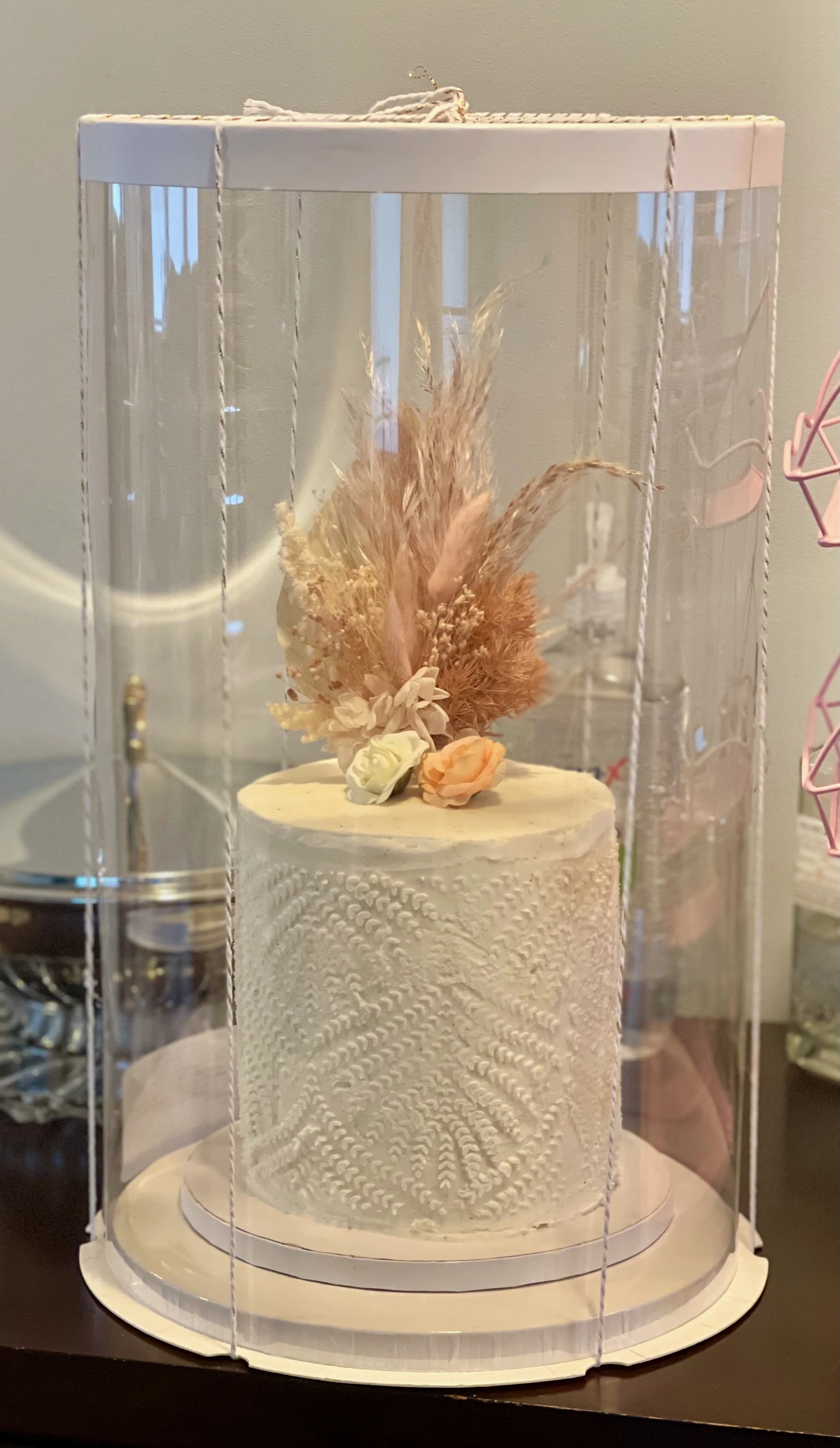 A white, decorated cake with textured icing and floral decoration on top, placed inside a clear plastic display case.