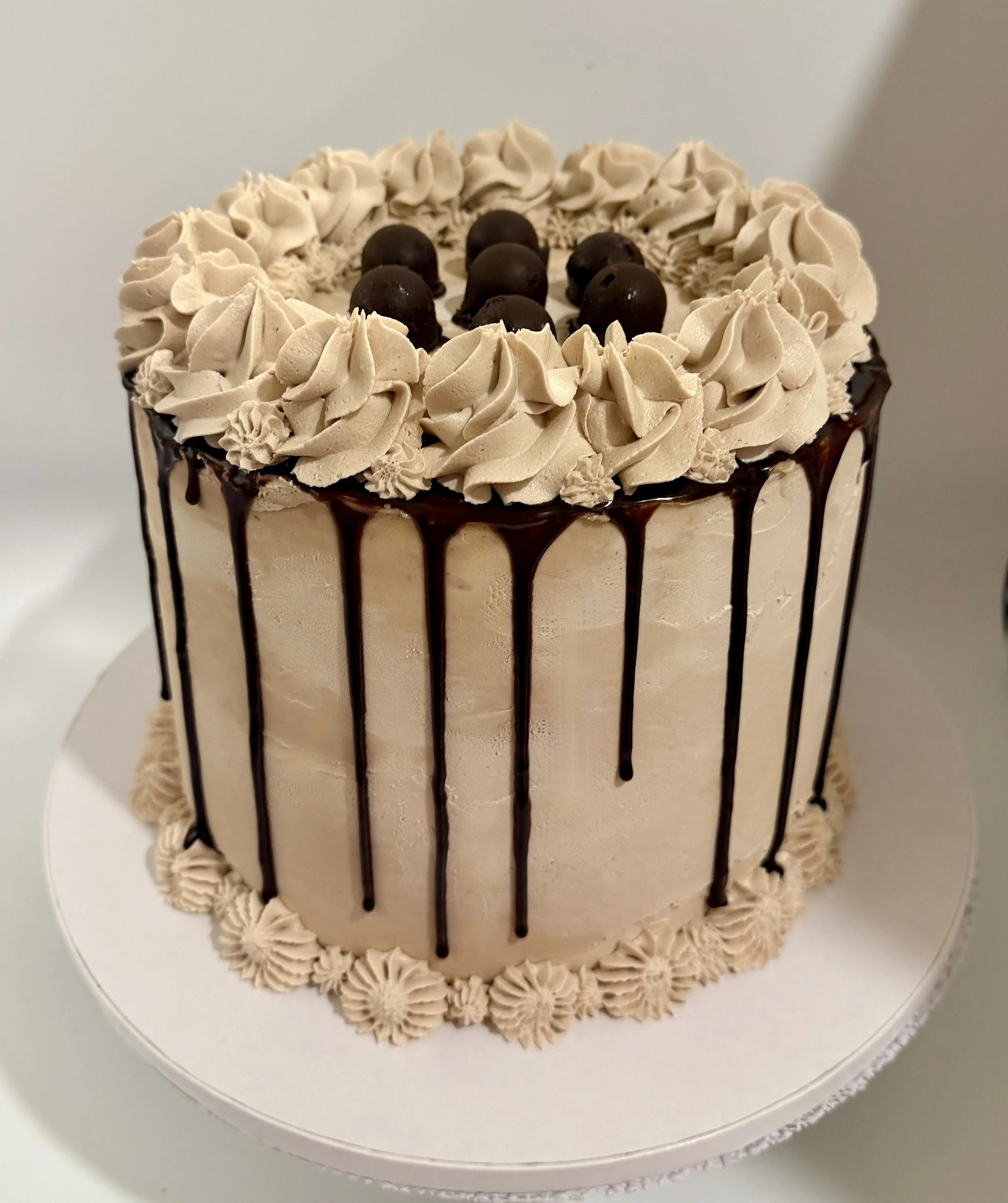 A round cake with chocolate frosting, chocolate drizzle hanging from the edges, and topped with chocolate truffles in the center. The cake has piped decorative frosting around the top and bottom edges.