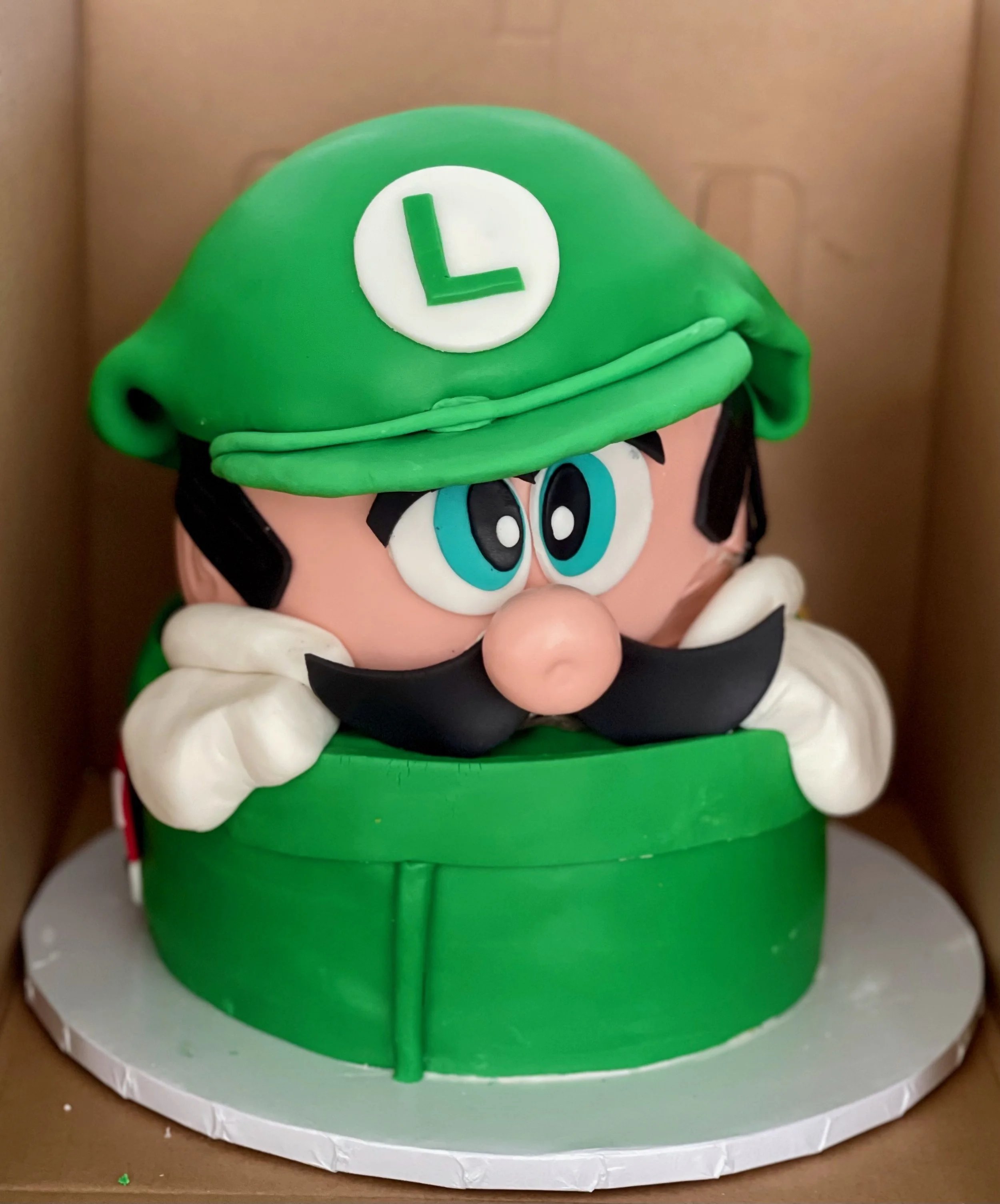 A cake decorated as Luigi from Nintendo with a green hat and white 'L' symbol, large eyes, and a black mustache.