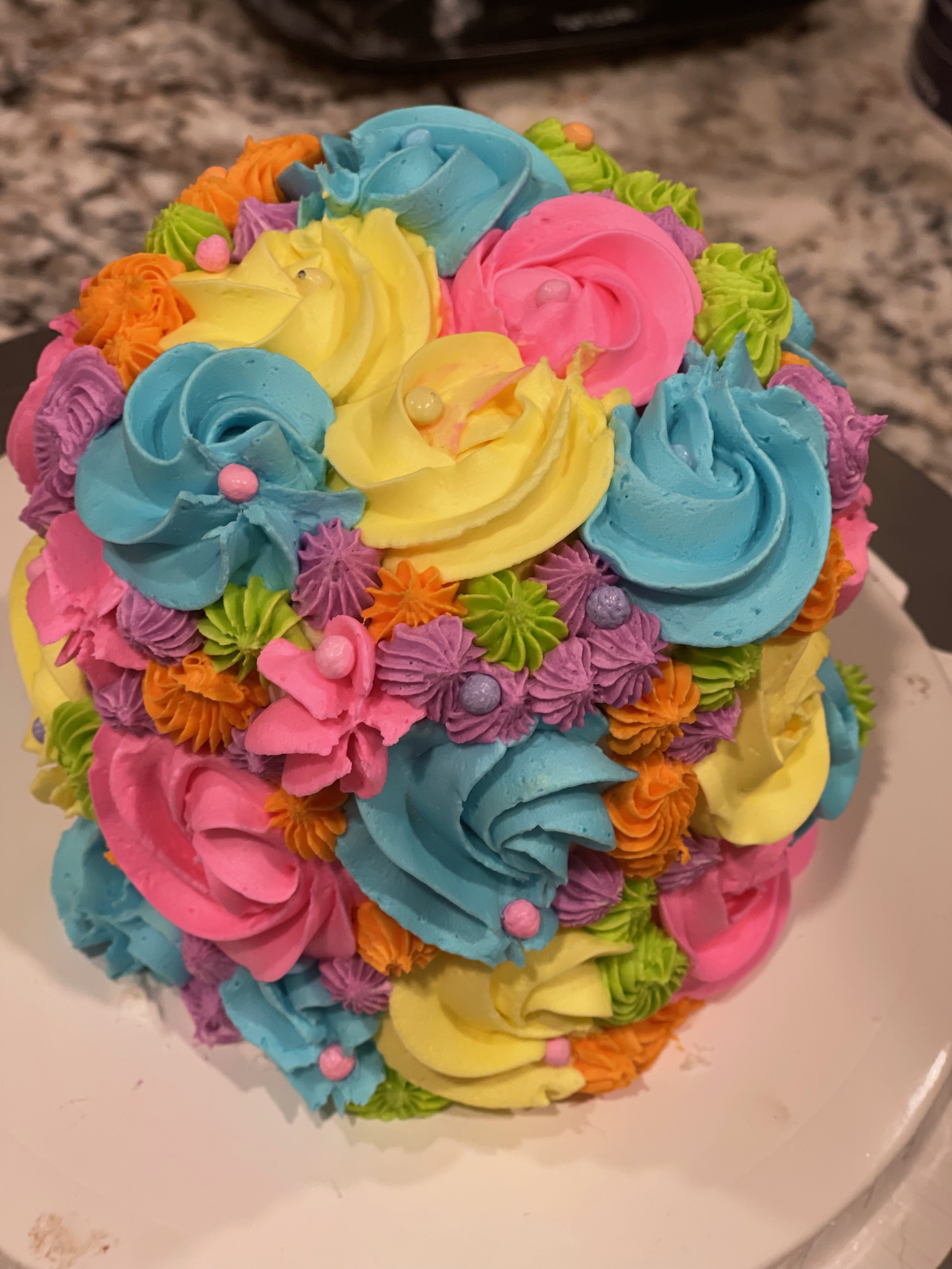 A cake decorated with colorful buttercream flowers in pink, blue, yellow, orange, green, and purple, with some small pearl-like edible decorations on top.