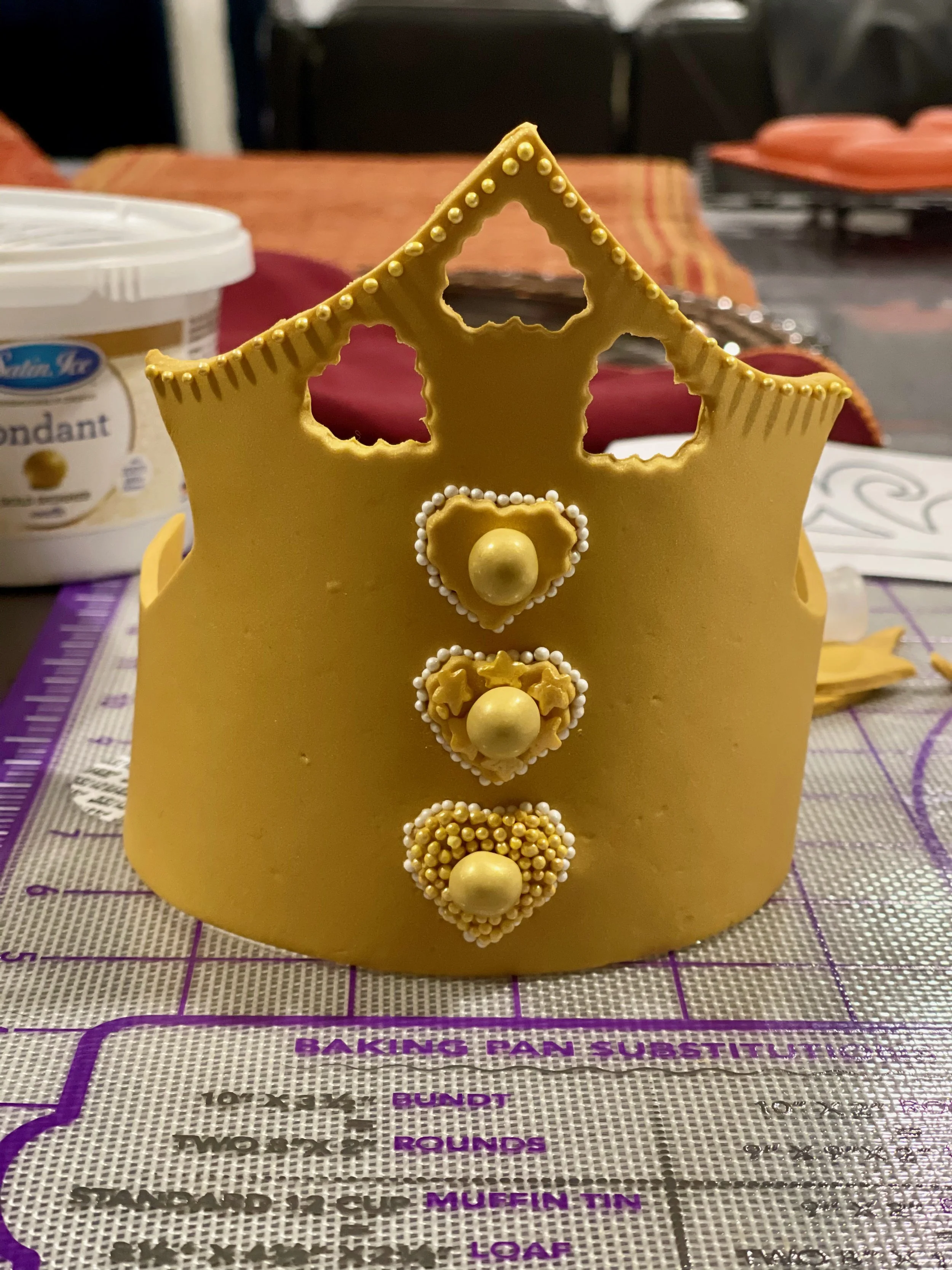 A gold fondant crown with sugar pearl details and heart and star cutouts, sitting on a silicone baking mat with purple baking instructions, with baking supplies in the background.
