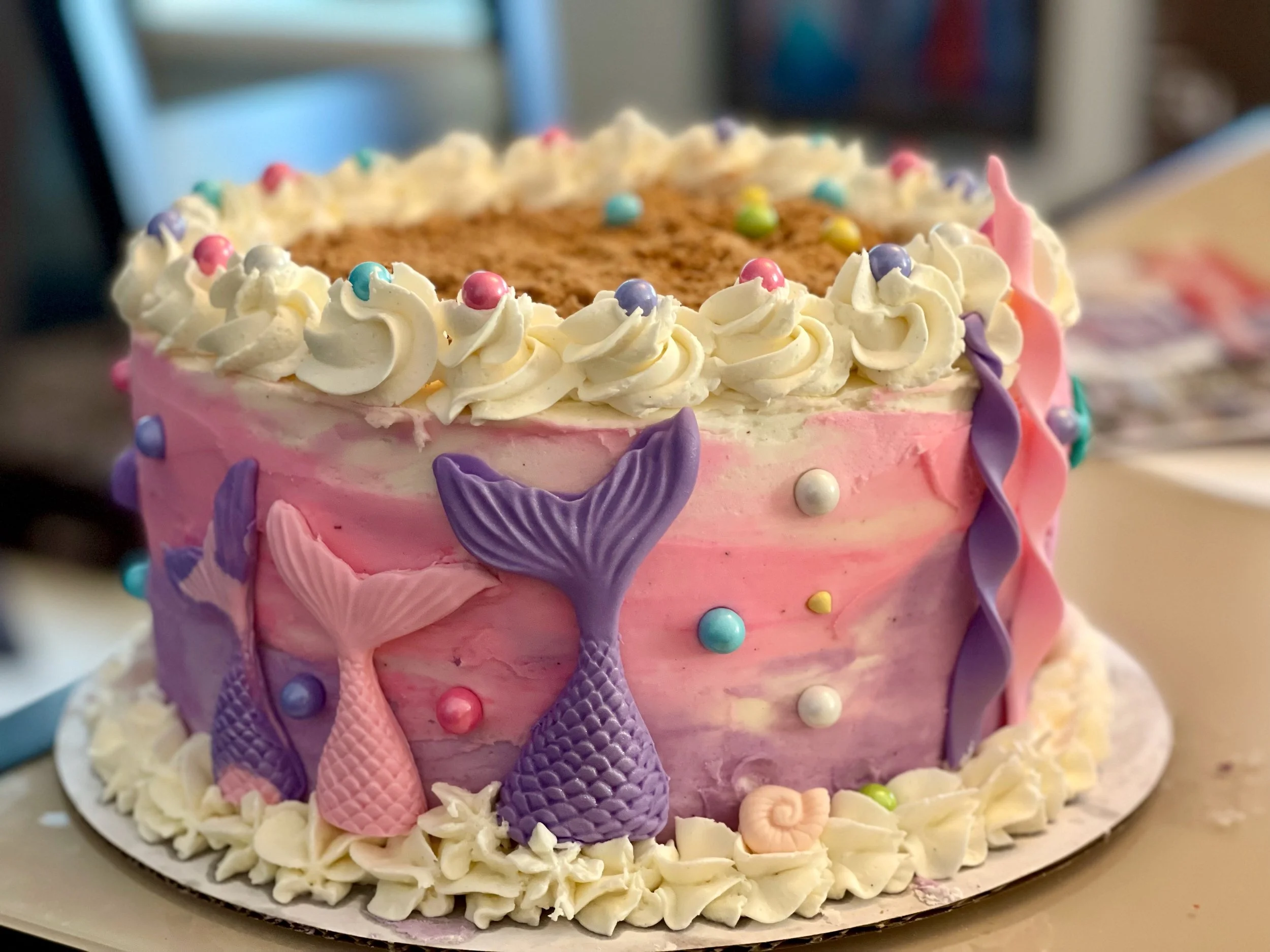 Colorful cake decorated with pastel-colored icing, mermaid tails, candies, and frosting, featuring a pink, purple, and white color scheme with frilled icing details.
