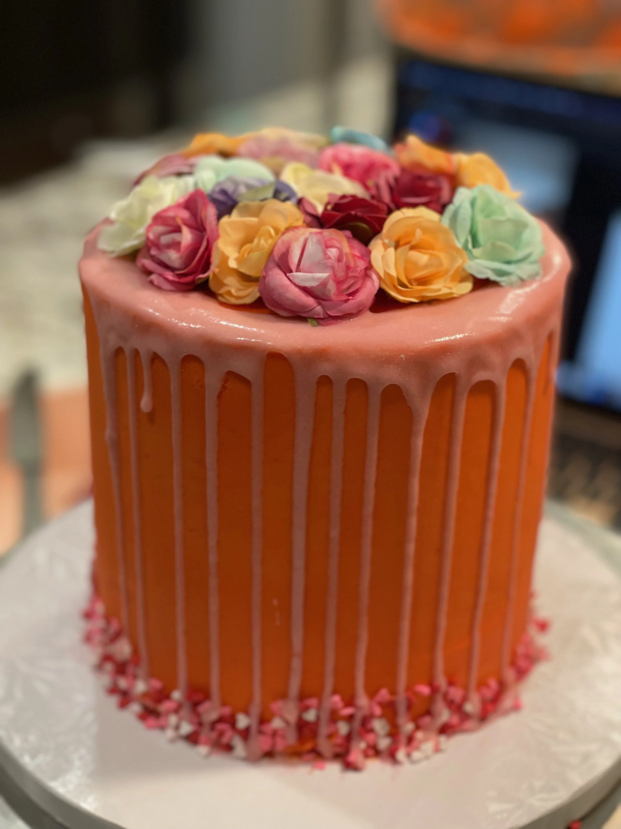 A round cake decorated with pink, orange, yellow, purple, and green silk roses on top, with white drip icing down the sides, and pink and white sprinkles around the base.