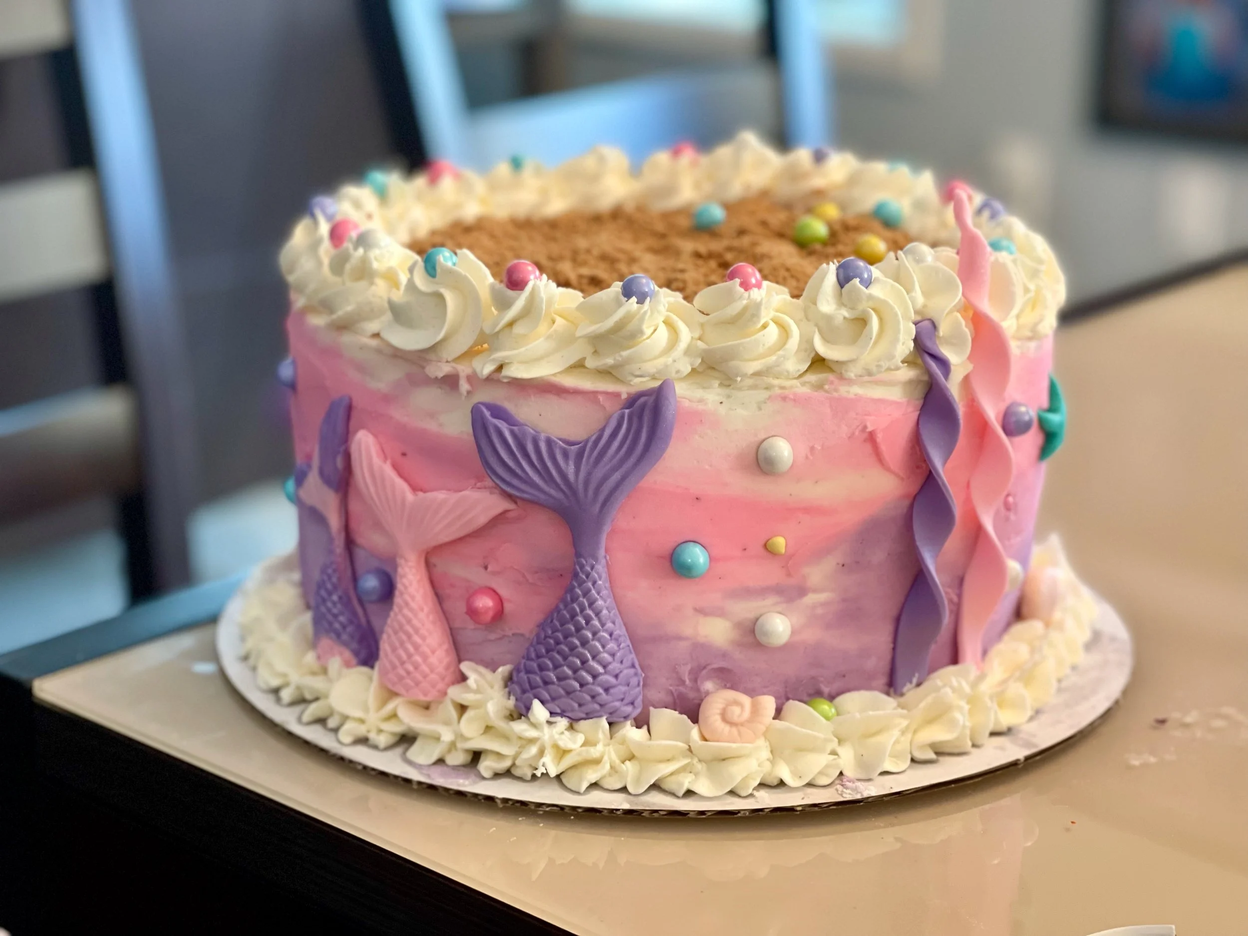 A pink birthday cake decorated with pastel-colored ice cream cone-shaped candies, sprinkles, and white frosting rosettes around the top edge. The cake has a striped pink and purple color theme with shell and pearl decorations.