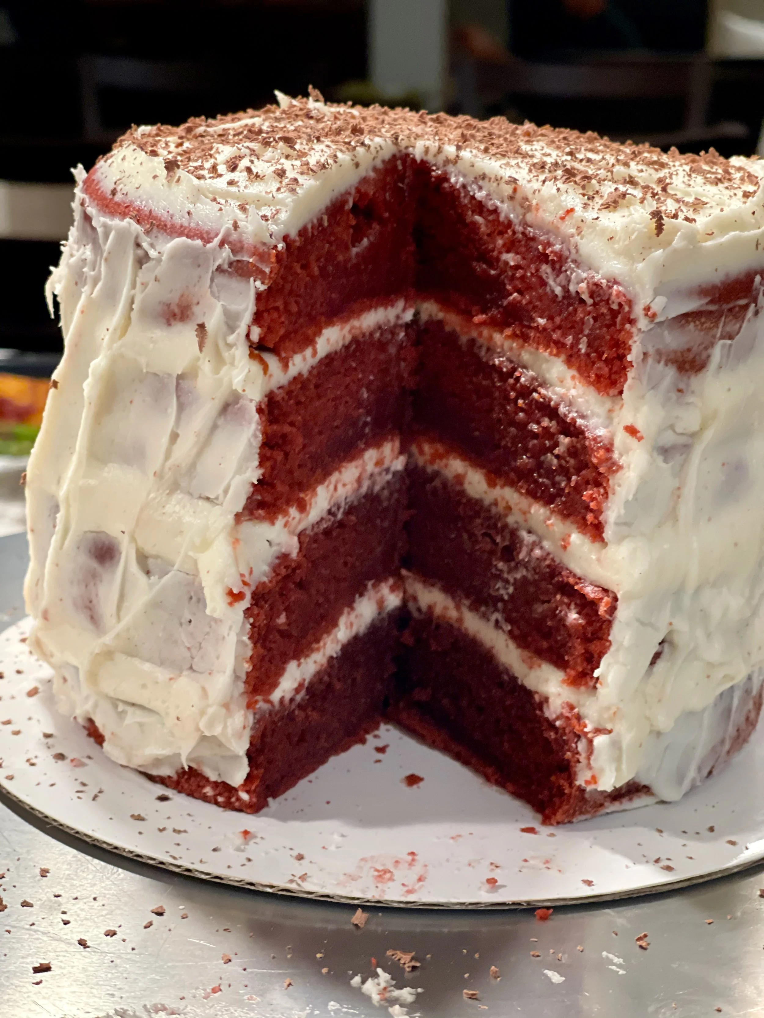 A partially sliced red velvet cake with cream cheese frosting and chocolate shavings on top, sitting on a white plate. Crumbs and frosting smears are visible around the cake.