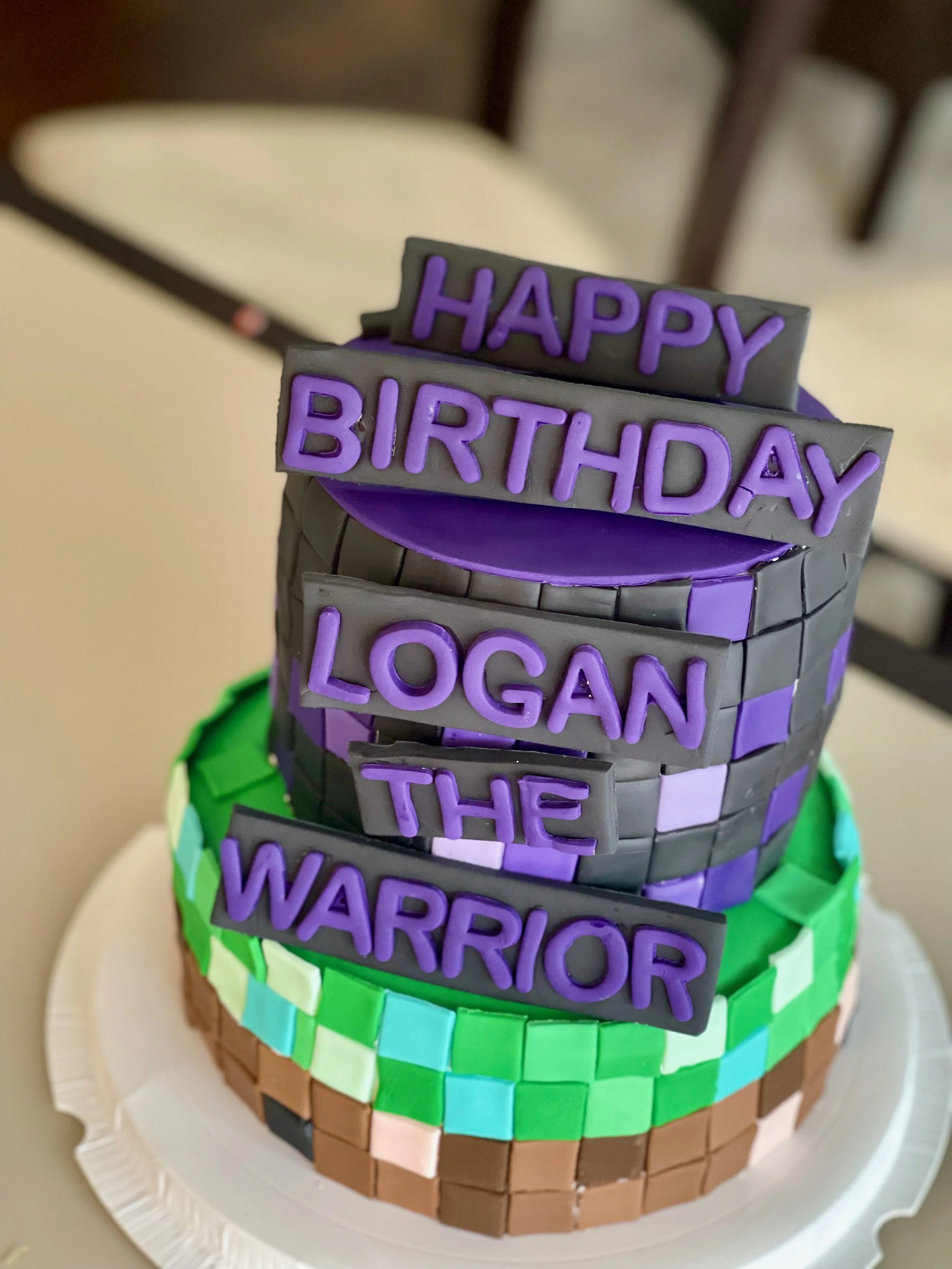 Colorful, tiered cake decorated with alphabet tiles in purple spelling out 'HAPPY BIRTHDAY'. The cake has a rainbow-colored tile border at the base and is placed on a white disposable plate.