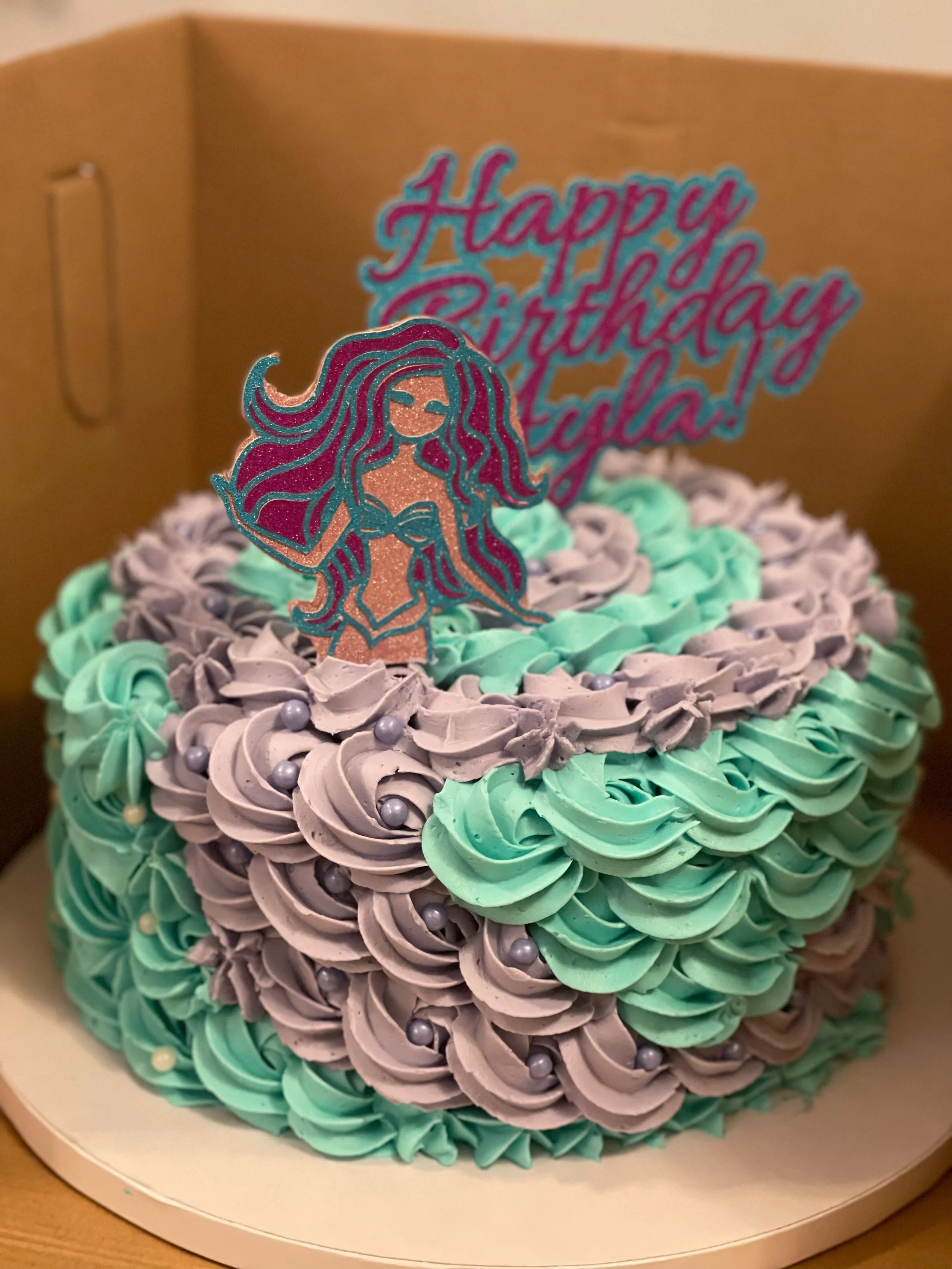 A birthday cake decorated with purple and mint green swirl frosting, silver edible pearls, and a topper that says 'Happy Birthday Edela!'. The cake is in a white box.