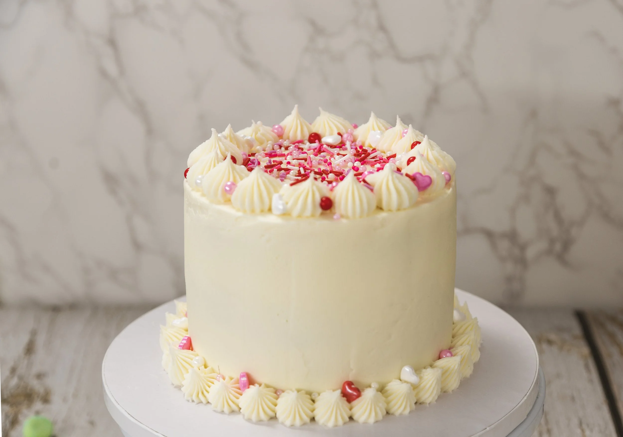 A ream-colored cake decorated with pink and white sprinkles, small pink hearts, and what appears to be piped frosting shells around the edges of each tier, placed on a white cake stand.