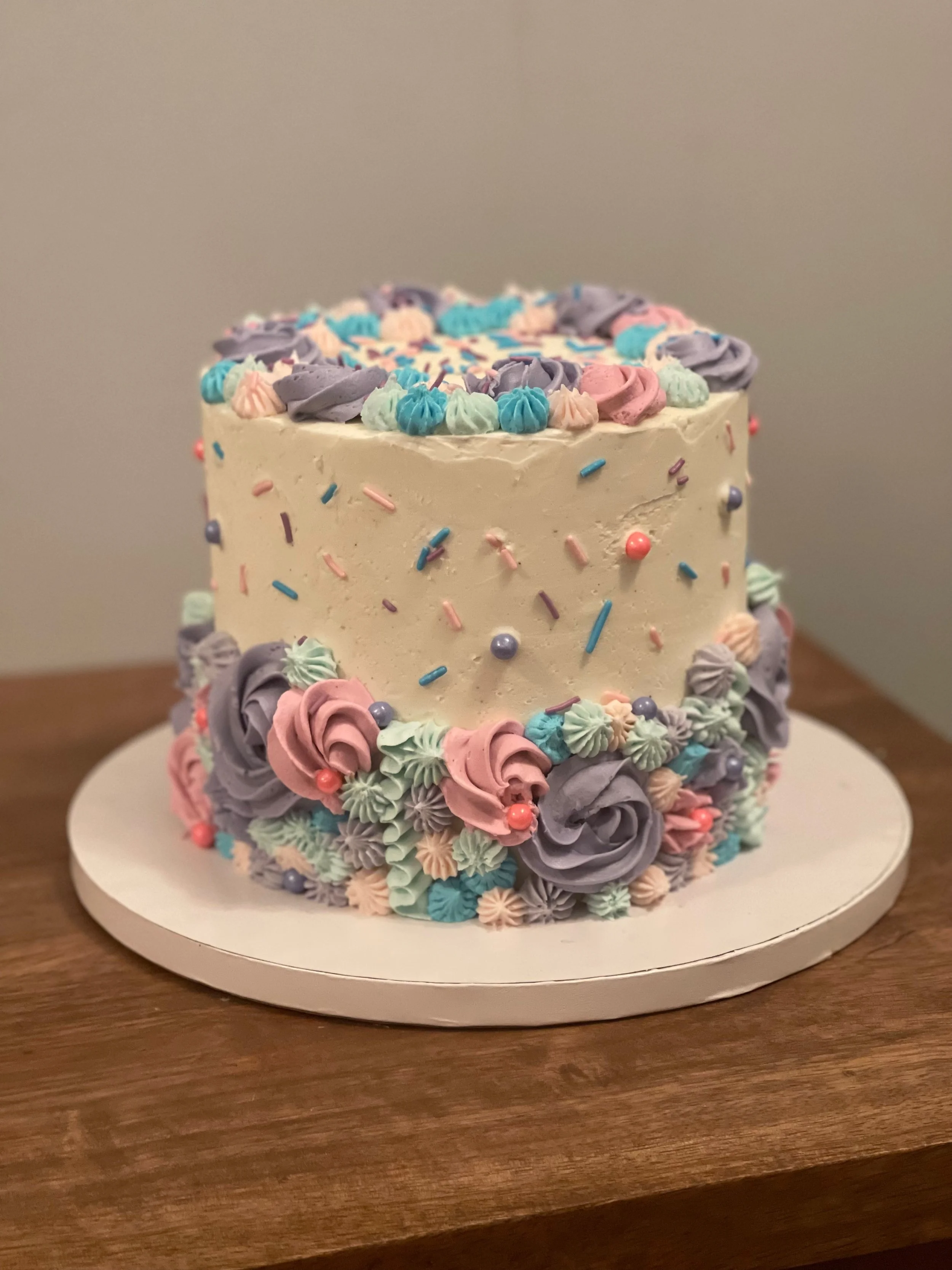 A birthday cake decorated with pastel-colored buttercream rosettes, sprinkles, and pearls, sitting on a white cake board on a wooden table.