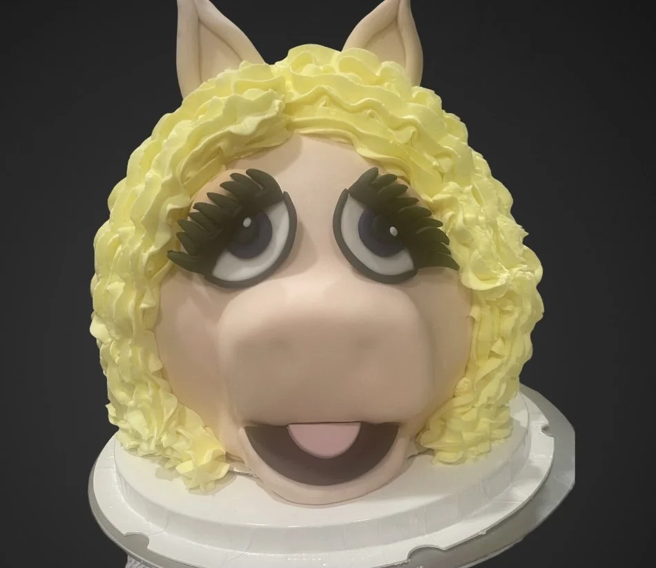 Cake decorated as Miss Piggy with blonde curly hair, big eyes with eyelashes, and ears, on a white cake stand.