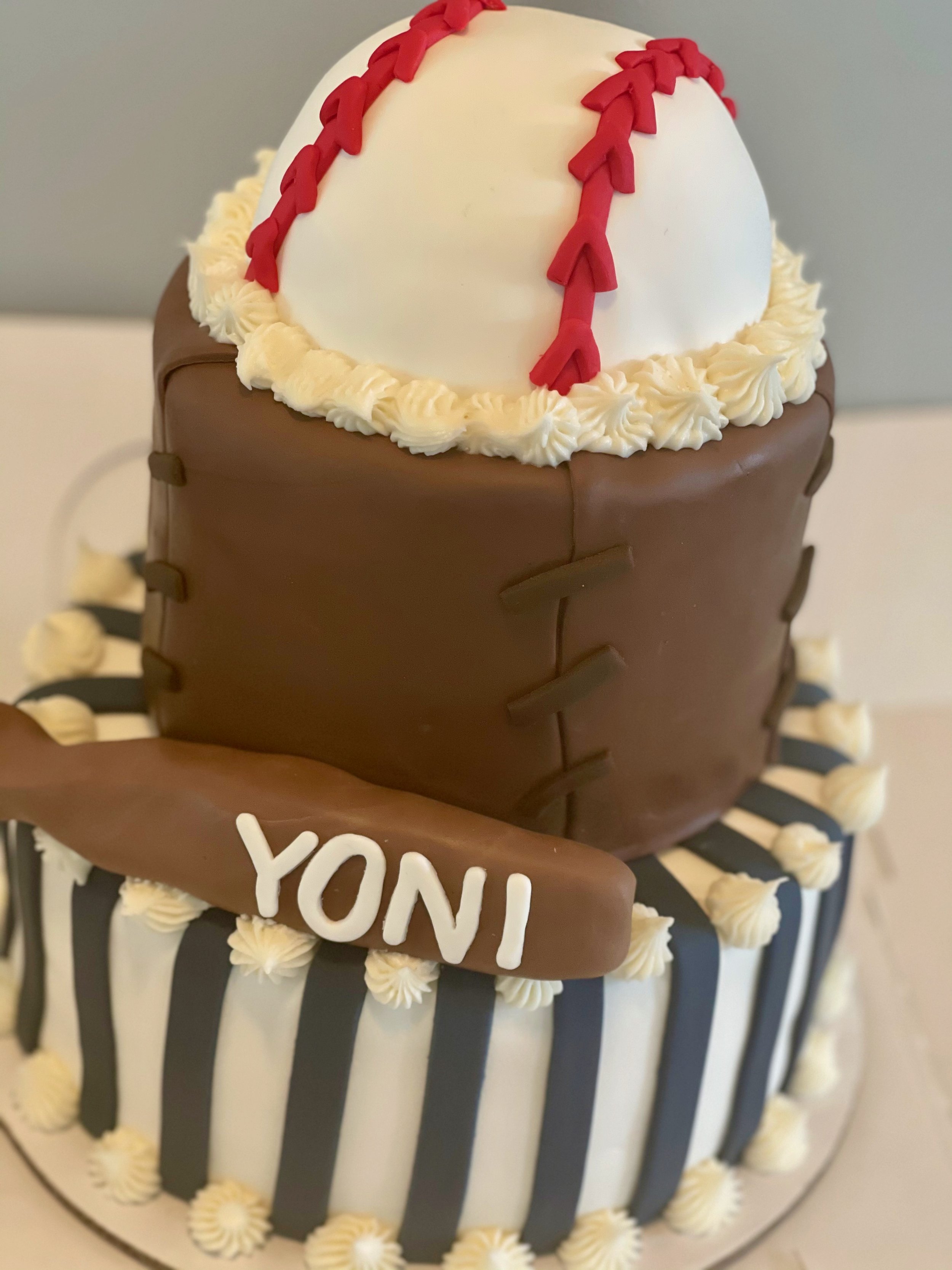 A round cake decorated to look like a baseball with white frosting, red stitching, and chocolate accents, featuring a chocolate plaque with the name 'YONI' in white letters.
