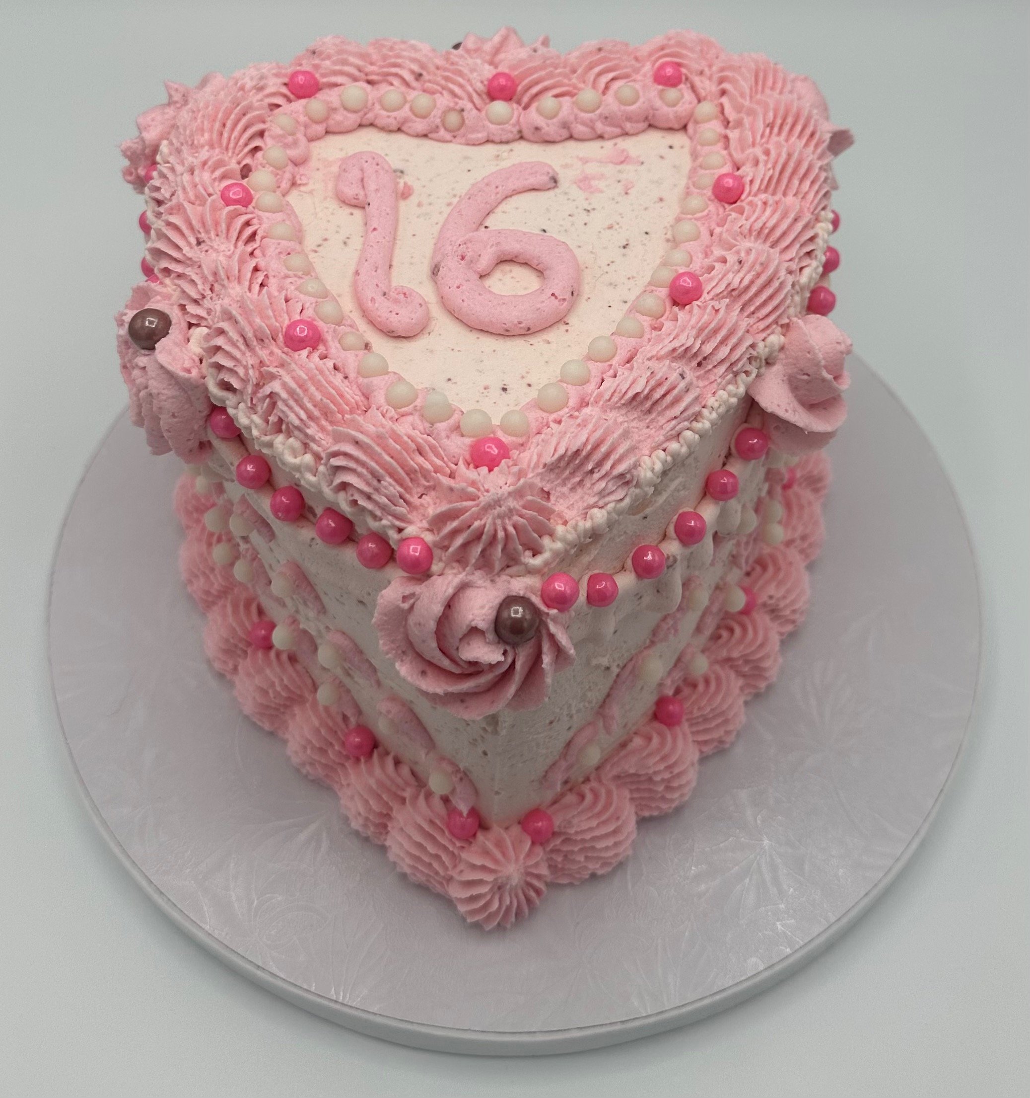 Heart-shaped birthday cake decorated with pink icing, white and pink round candy accents, and the number 16 in pink icing on top, placed on a white textured cake board.