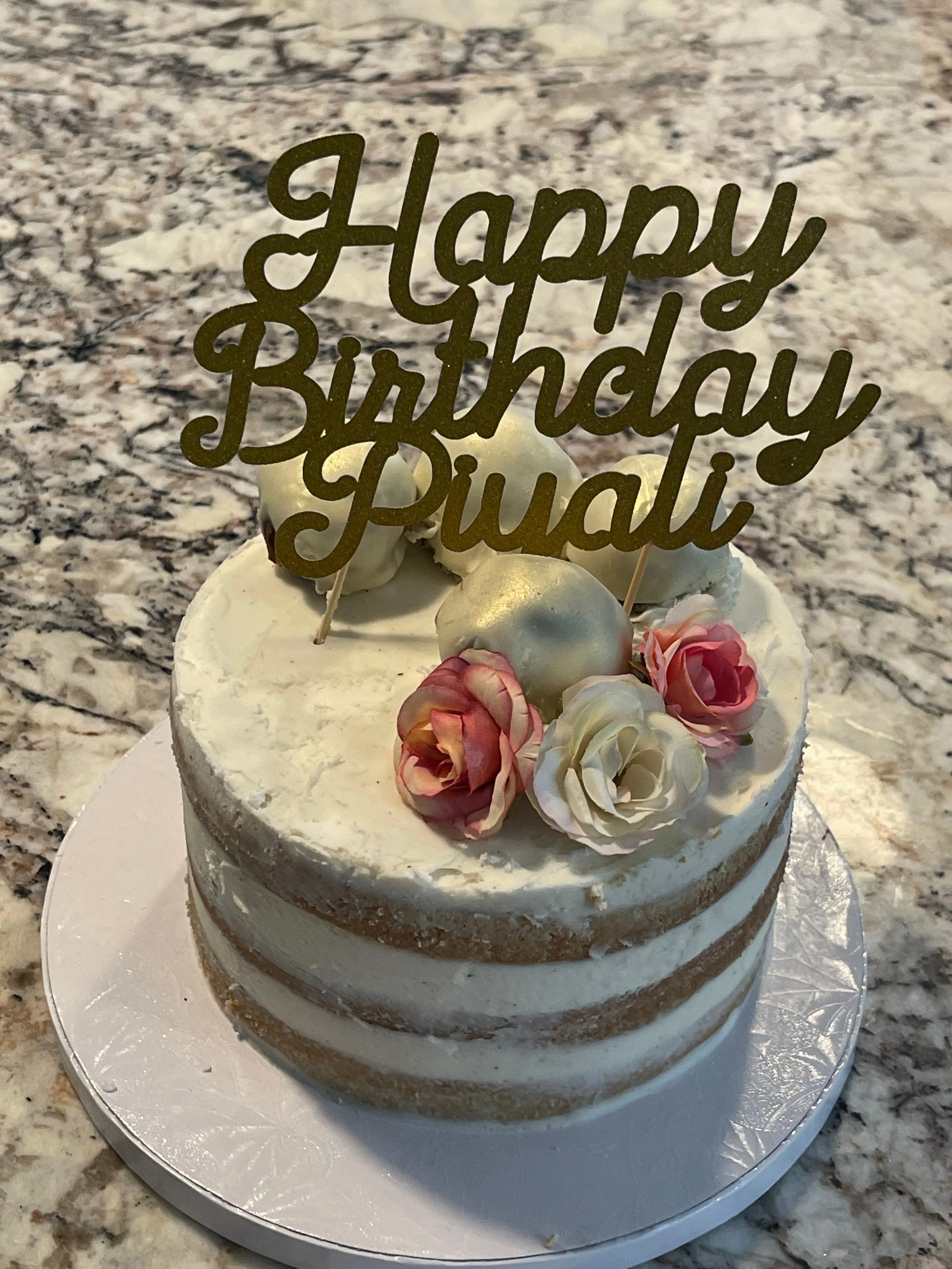 A white birthday cake decorated with pink and white flowers and topped with a gold 'Happy Birthday' cake topper on a marble countertop.
