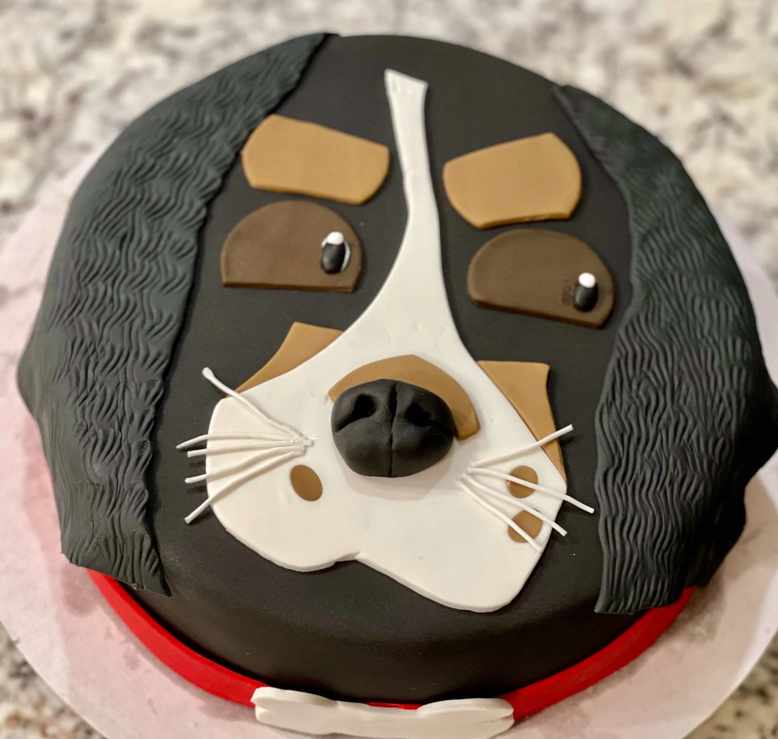 Close-up image of a decorated cake featuring a stylized dog ace with black, white, brown, and beige fondant elements, set on a black and red cake board.