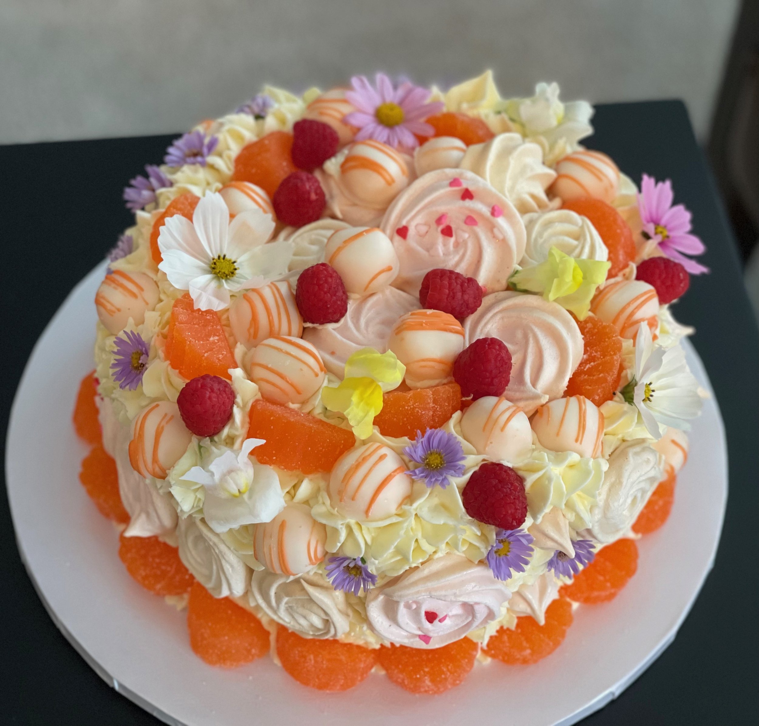A colorful cake decorated with raspberries, candy orange slices, small white meringue cookies, edible flowers, and pink heart-shaped sprinkles.