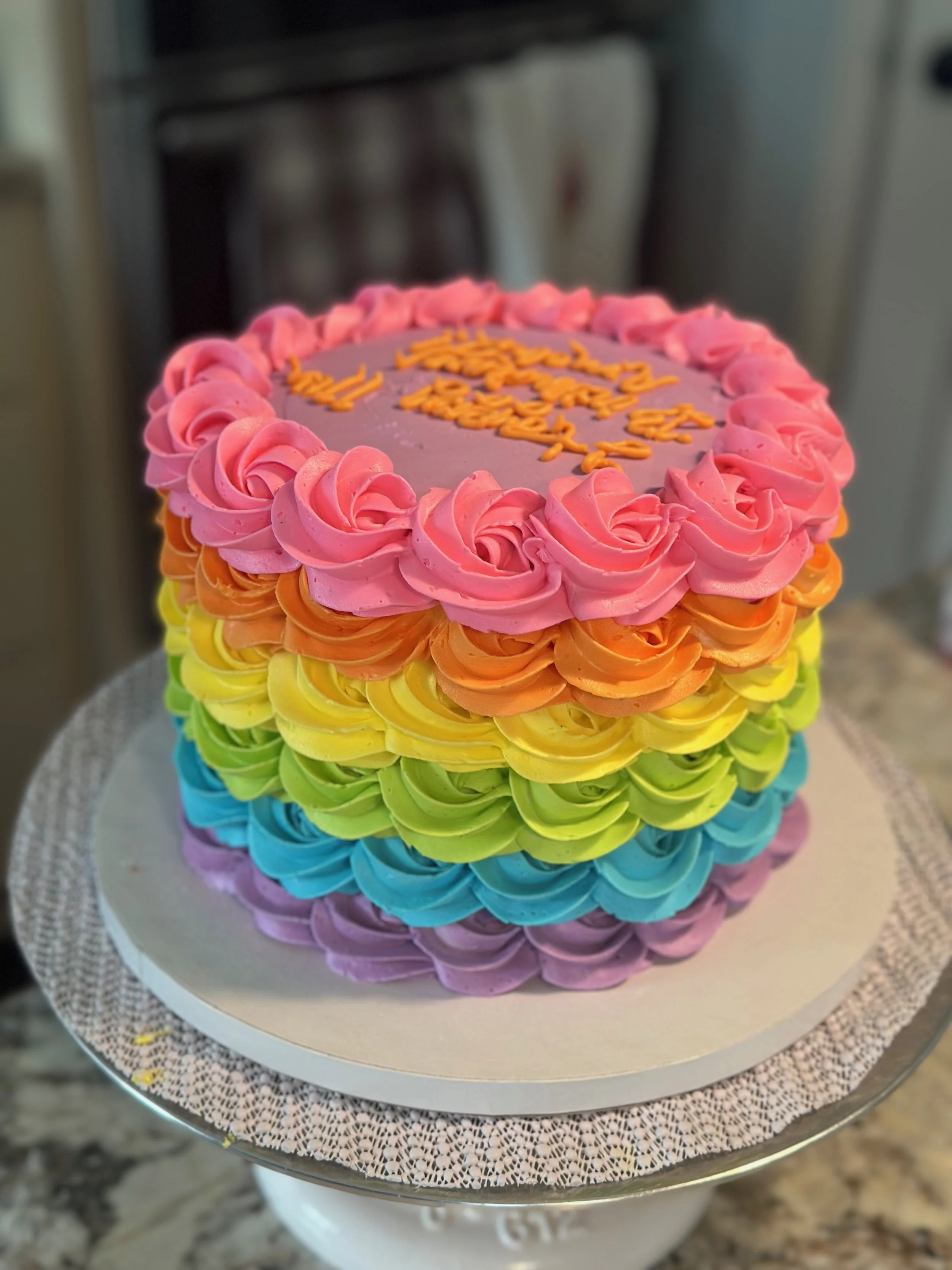 A rainbow-colored layered cake decorated with pink, orange, yellow, green, blue, and purple frosting roses on a silver cake stand.