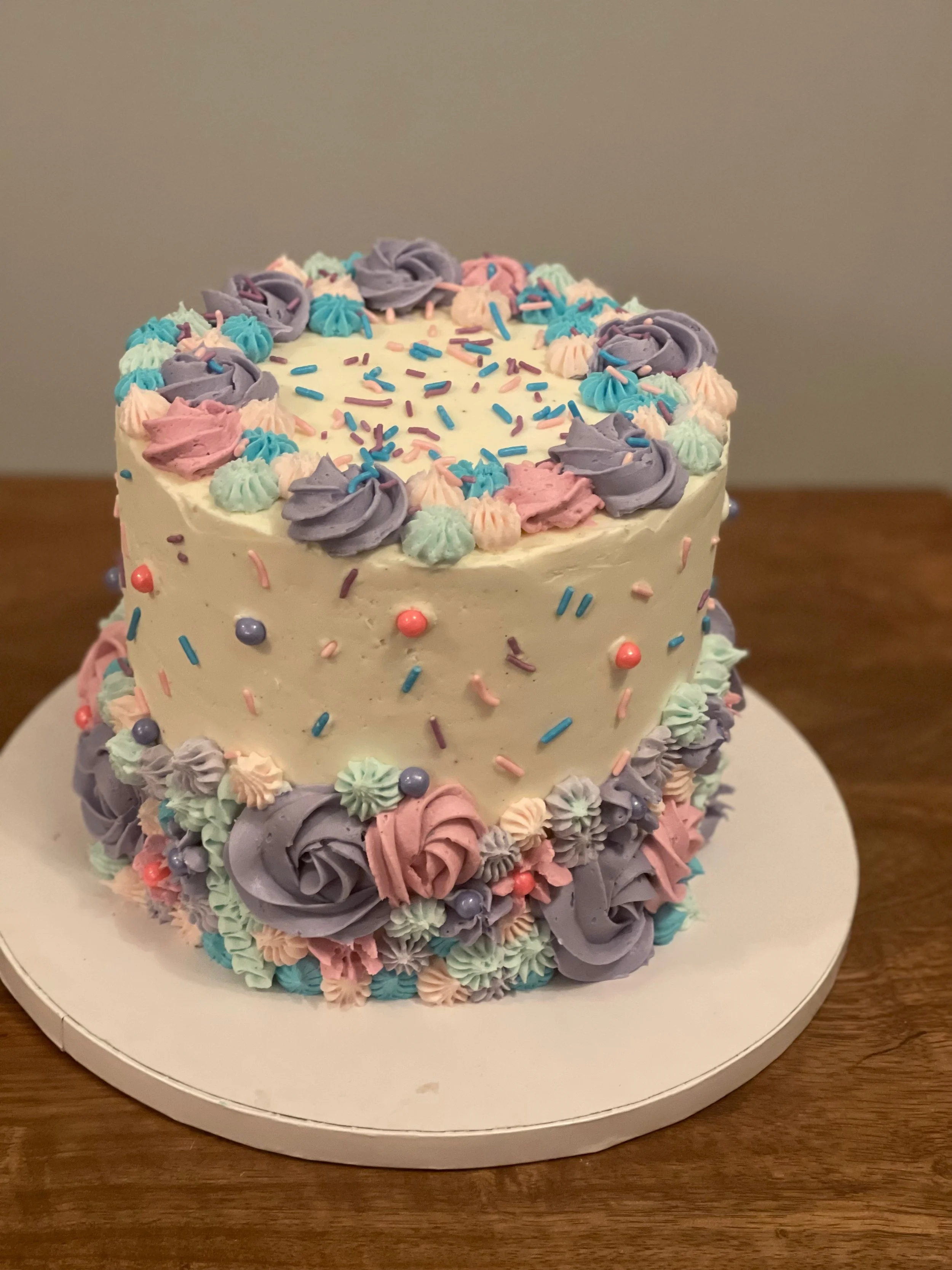 A birthday cake decorated with purple, pink, and mint green buttercream flowers and sprinkles, placed on a white cake stand on a wooden table.