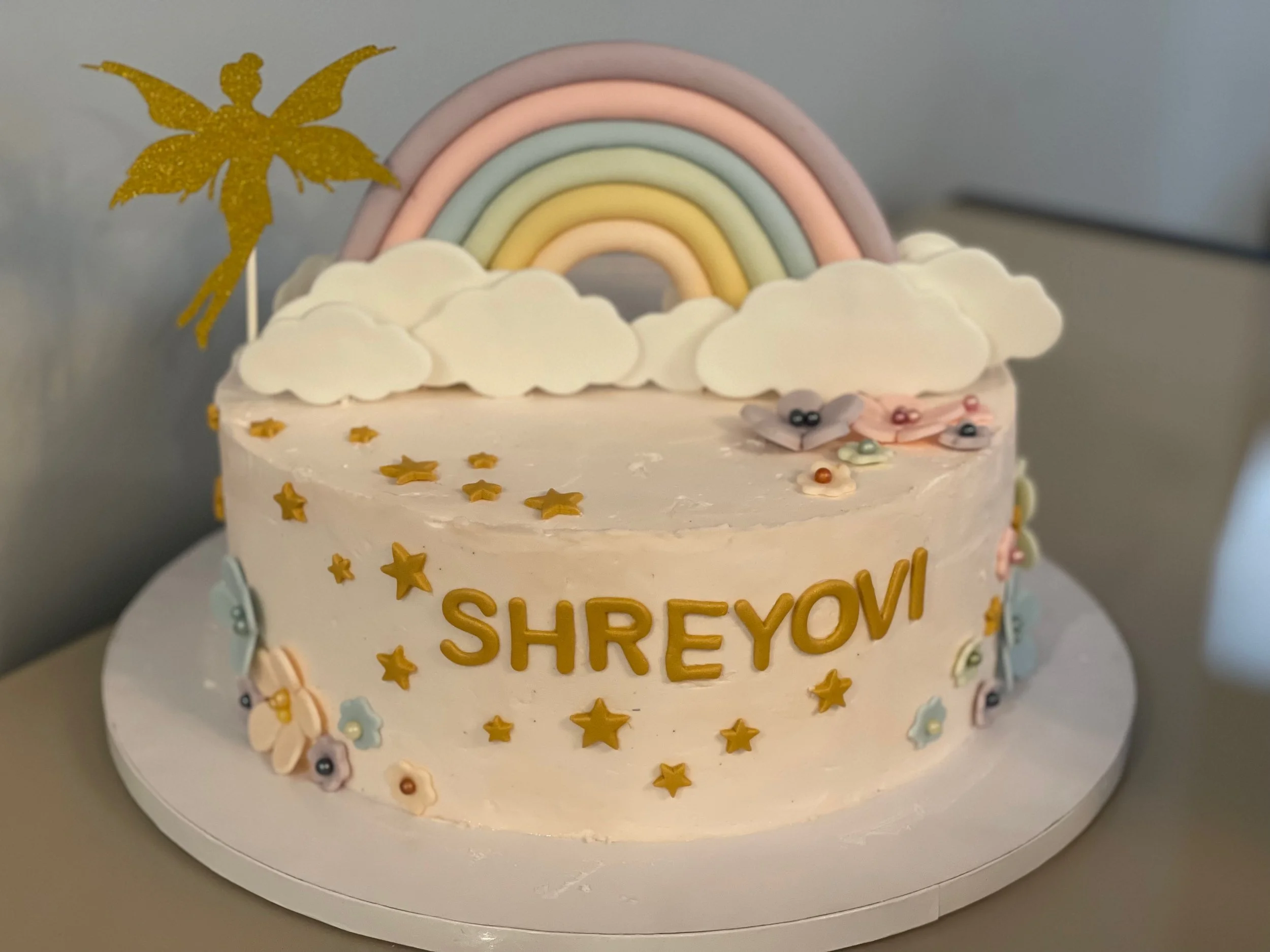 A birthday cake decorated with a rainbow, clouds, and stars, with golden glittery decorations, and the name 'Shreyov' written on it.