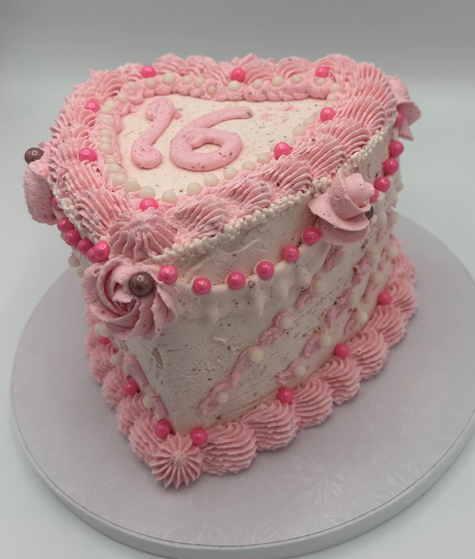Pink heart-shaped cake decorated with pink and white icing, pink and white pearls, and the number 16 in pink icing on top. The cake has piped pink icing around the edges and is displayed on a silver cake board.
