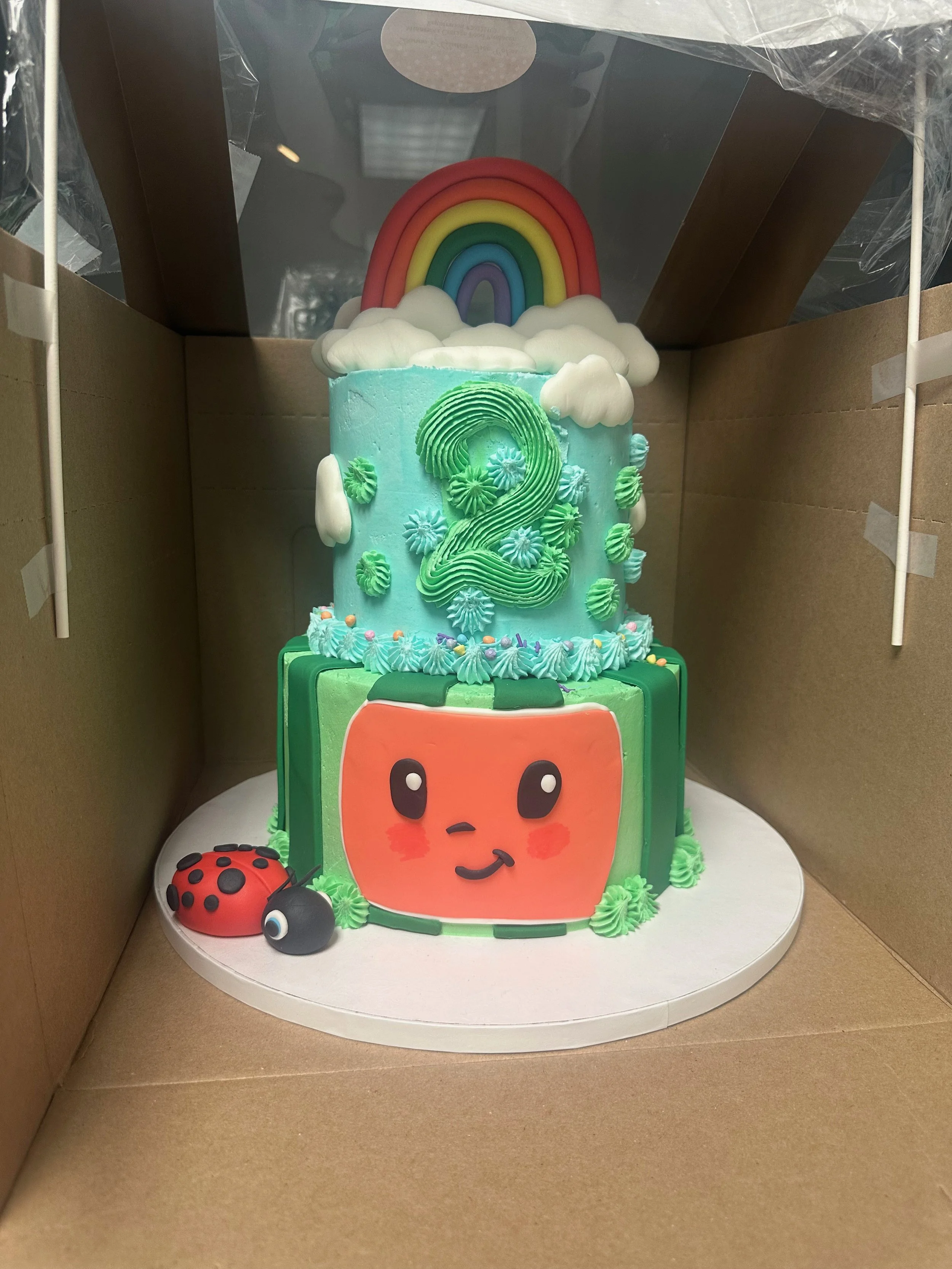 Decorated cake with a watermelon face, surrounded by green and blue frosting, featuring a smiling face with blush cheeks. On top, there is a rainbow with clouds. The cake is inside a cardboard box with a clear plastic window on top.