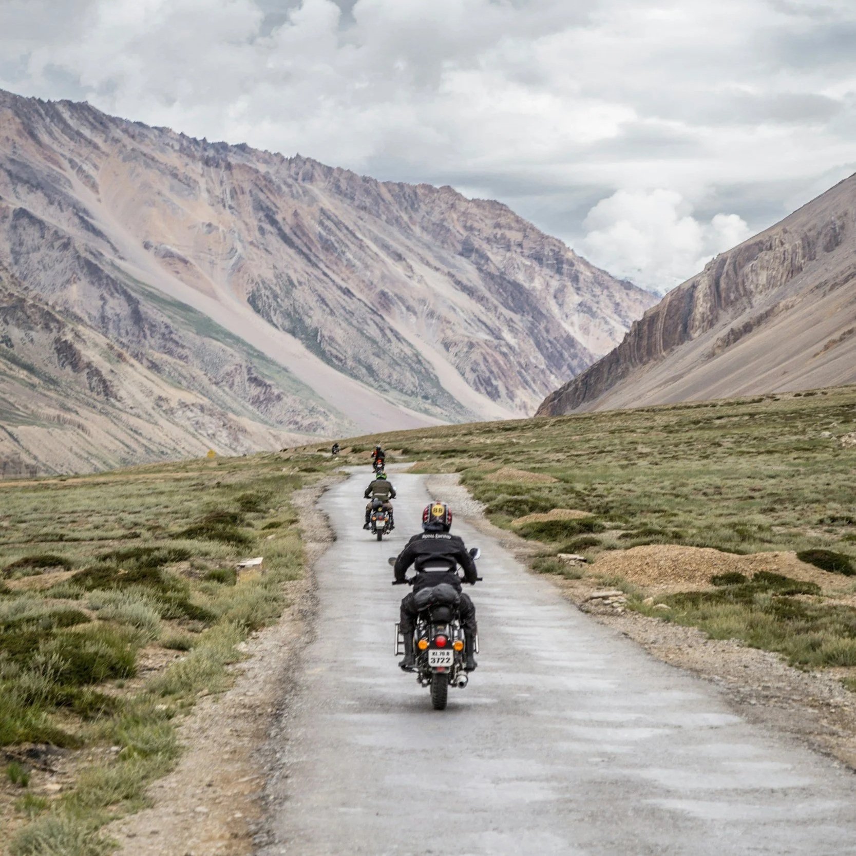 Group of motorcyclists riding on a scenic mountain road with rugged terrain and cloudy sky in the background.