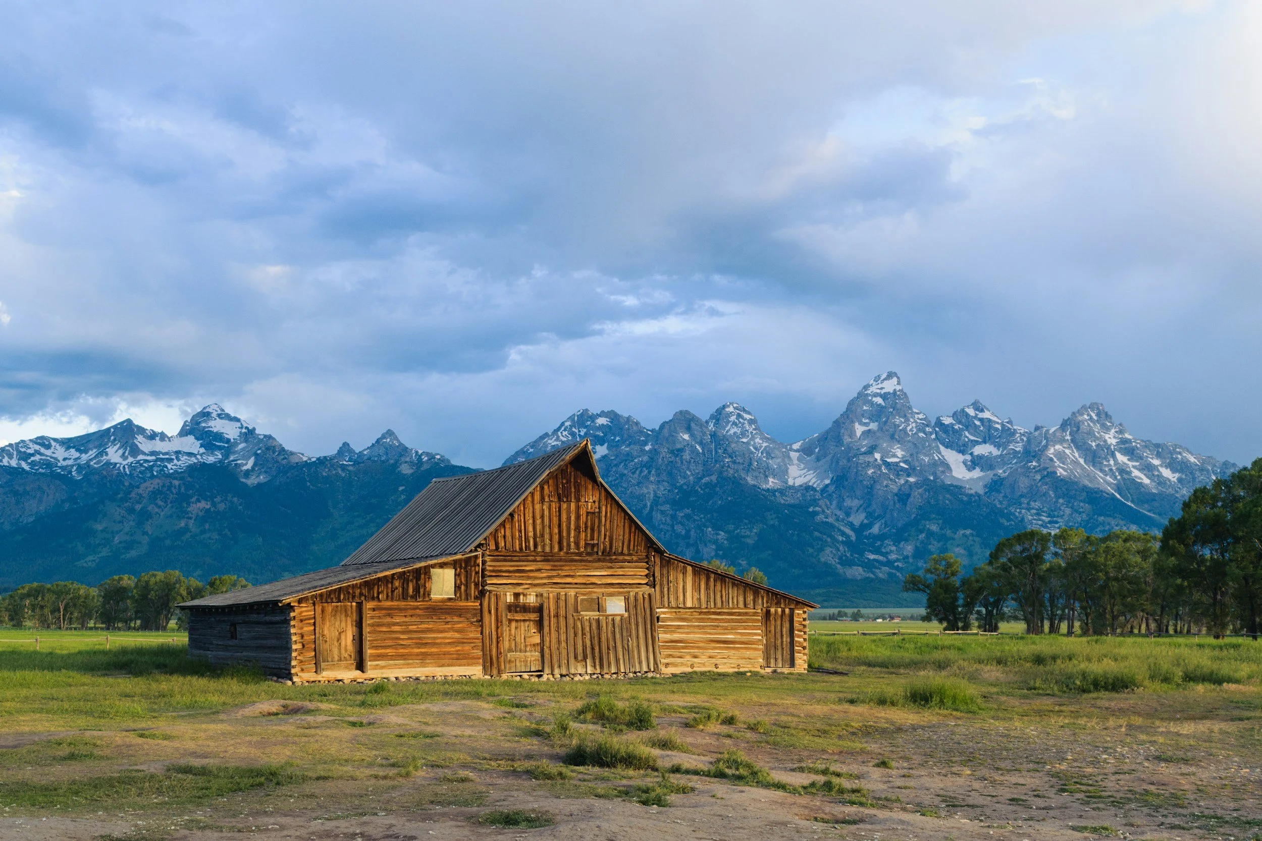 A rustic wooden barn in a green field with a mountain range and partly cloudy sky in the background.