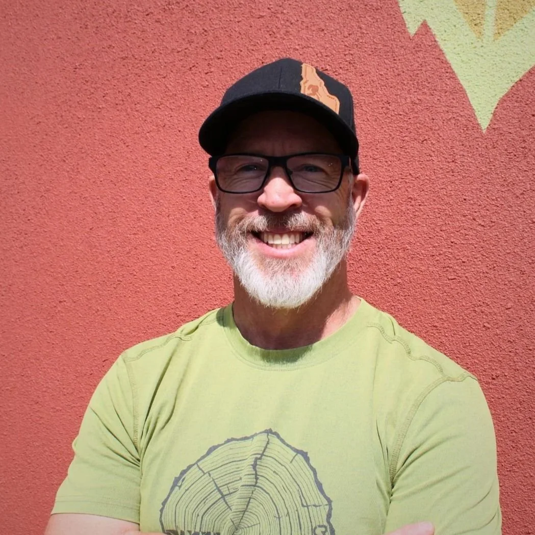 Smiling man with glasses, a beard, and a mustache wearing a black cap and a yellow T-shirt with a tree ring design, standing in front of a red stucco wall with tree canopy shadow.
