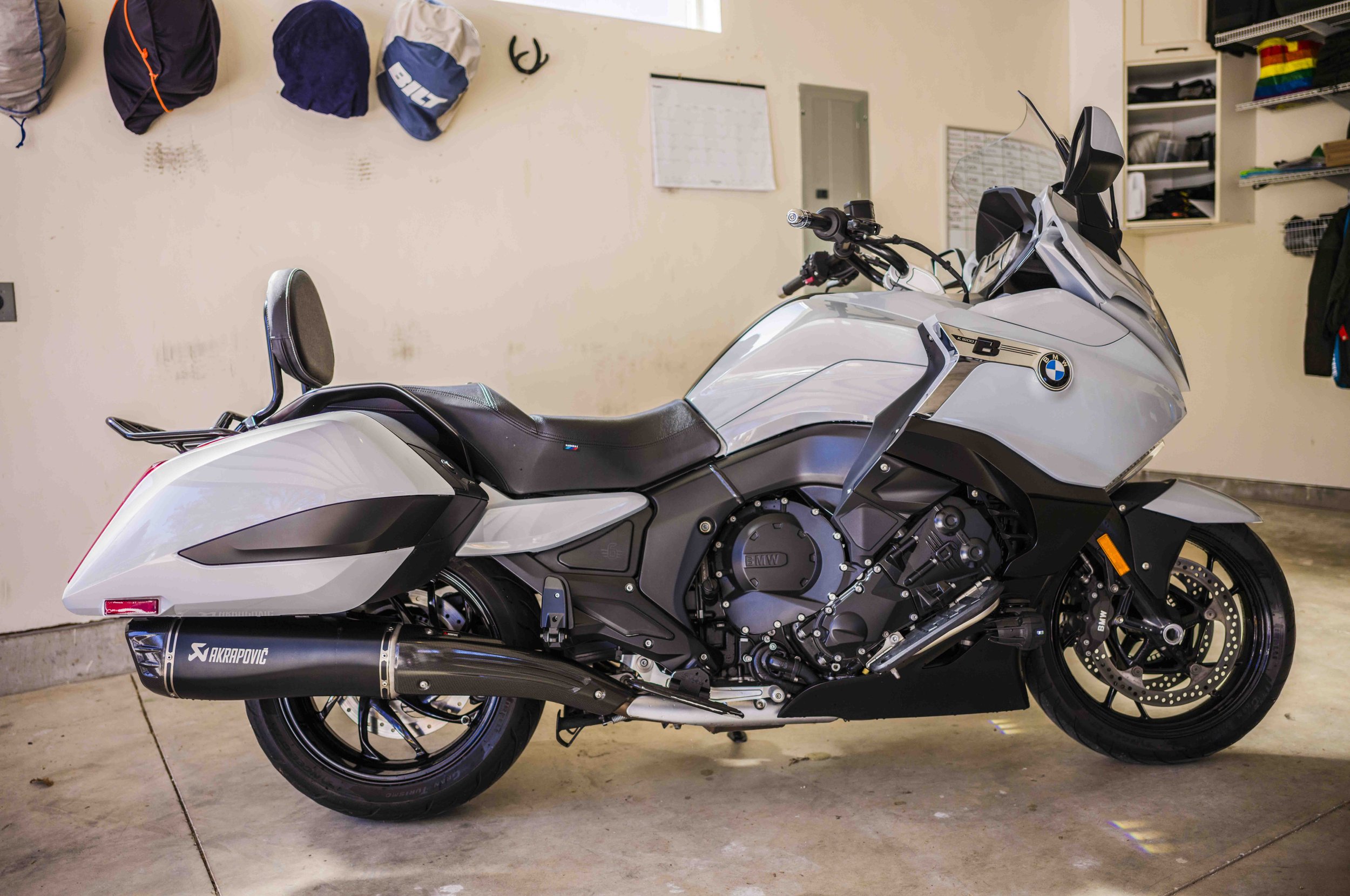 A white BMW touring motorcycle with black accents, parked inside a garage, featuring an Akrapovic exhaust and a small backrest for a passenger.