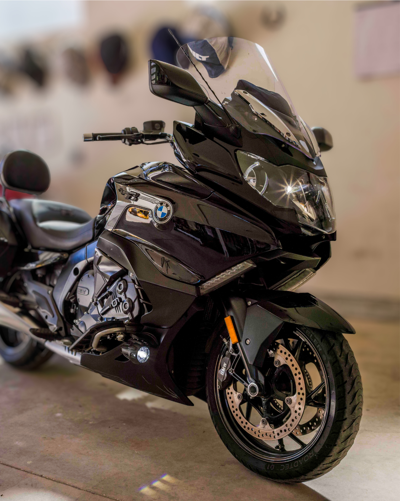 A black BMW motorcycle parked indoors on a concrete floor with a beige wall in the background. The motorcycle has side saddlebags and a windshield.