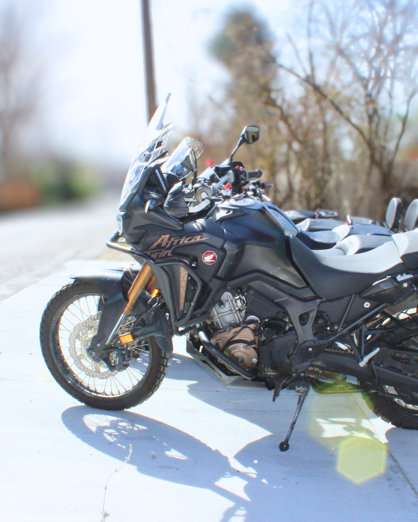 Black Honda Africa Twin touring motorcycle parked on a sidewalk with other motorcycles behind it, reflected sunlight, some trees in the background.
