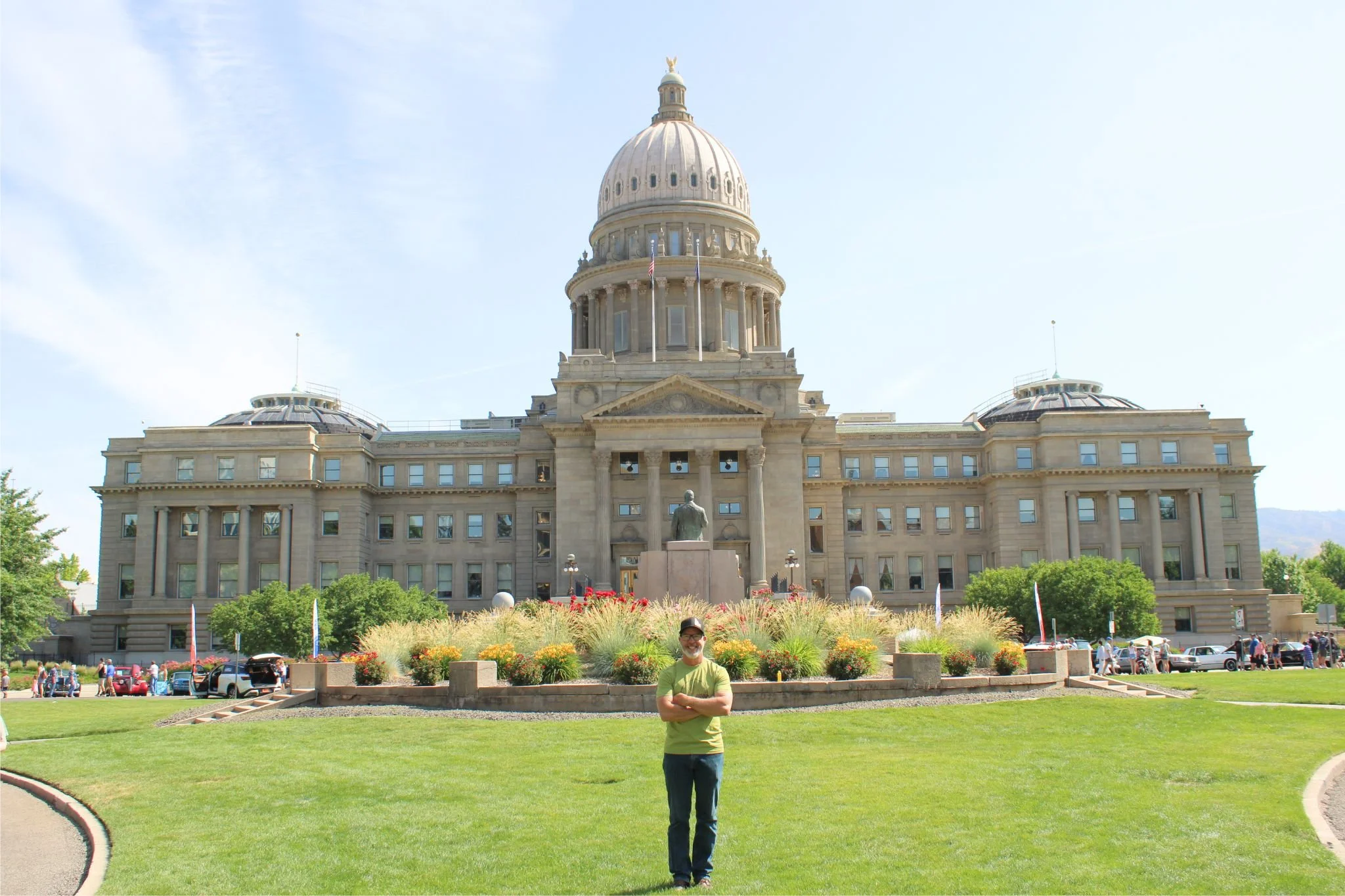 A man in a green t-shirt and jeans standing with arms crossed in front of the Utah State Capitol building on a sunny day.